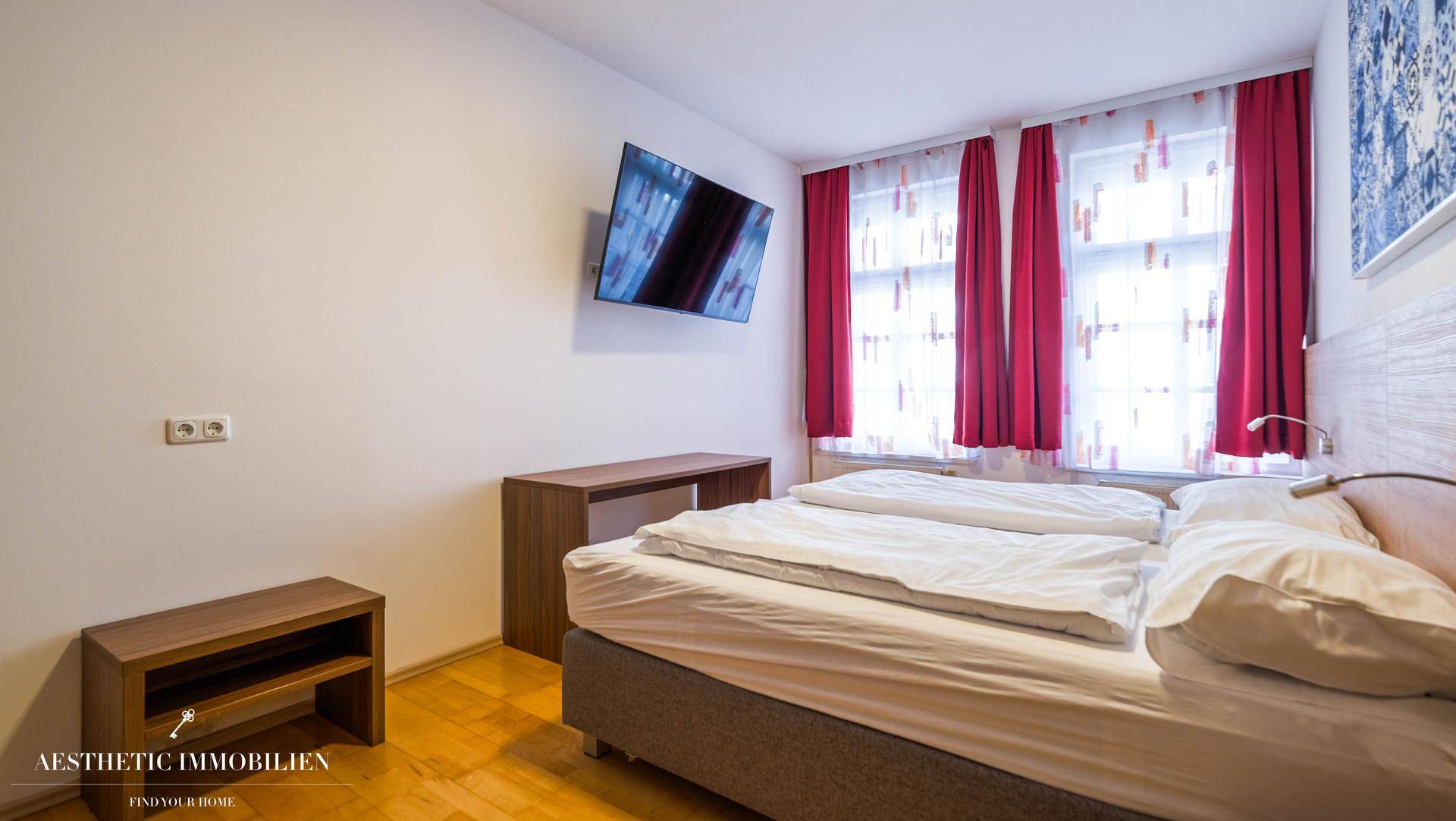 Schlafzimmer mit zwei Betten, Holzboden und Fenstern mit roten Vorhängen.