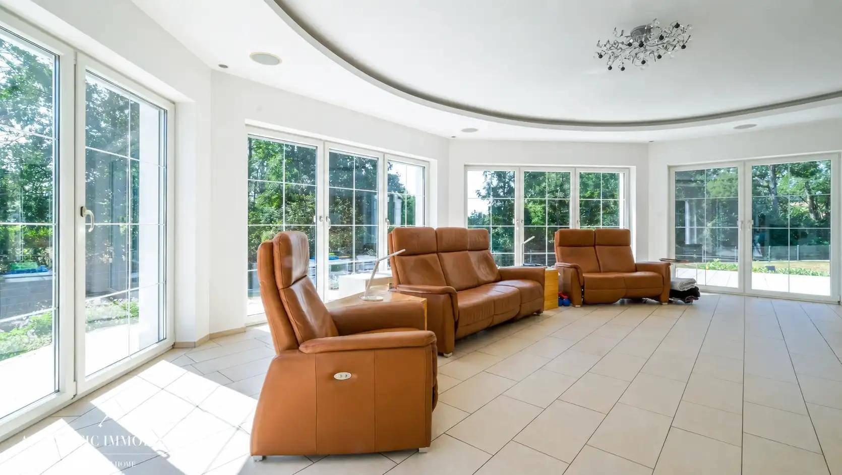 Helles Wohnzimmer mit großen Fenstern, Fliesenboden und bequemen Ledersofas mit Gartenblick.