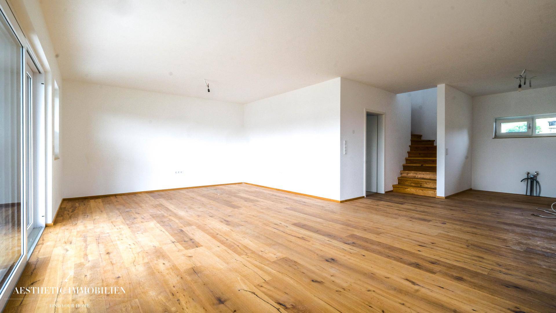 Geräumiger, leerer Wohnbereich mit Holzboden, Treppe und Zugang zum Außenbereich.