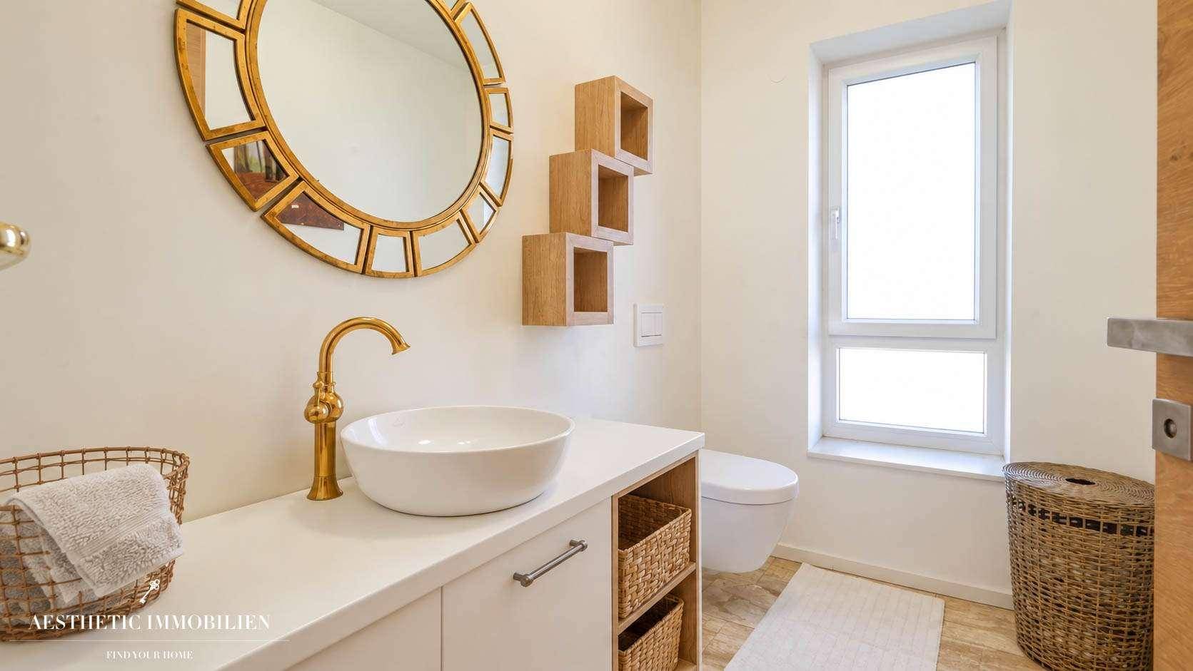 Modernes Gäste-WC mit rundem Spiegel, goldenem Wasserhahn und Fenster.
