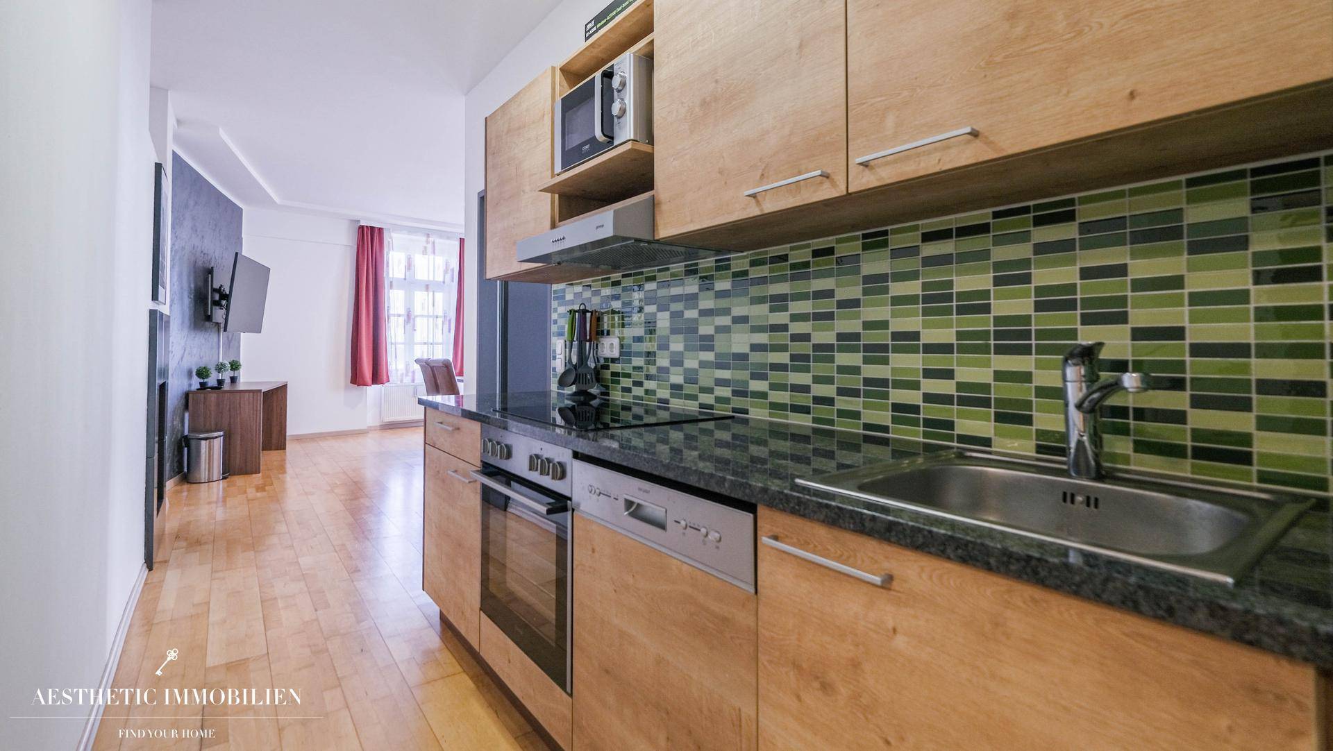 Funktionale Küche mit Holzschränken, grünen Mosaikfliesen und Blick in den Essbereich.