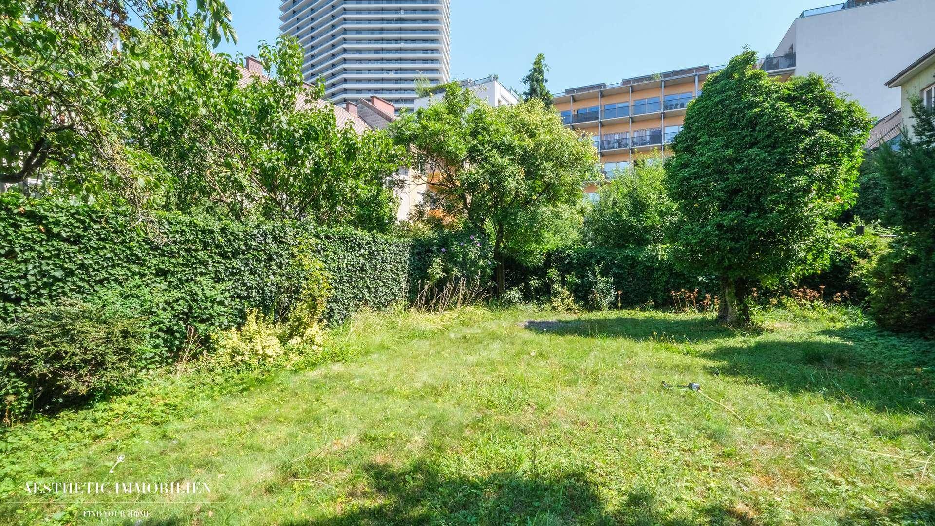 Grüner Gartenbereich mit Rasenfläche, Hecke und Blick auf die Stadtbebauung.