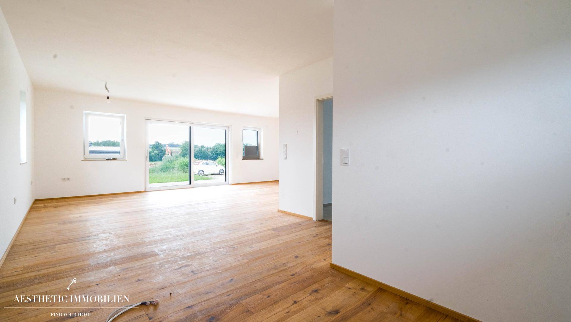 Heller, leerer Wohnbereich mit Holzboden und großen Fenstern, die viel Tageslicht hereinlassen.