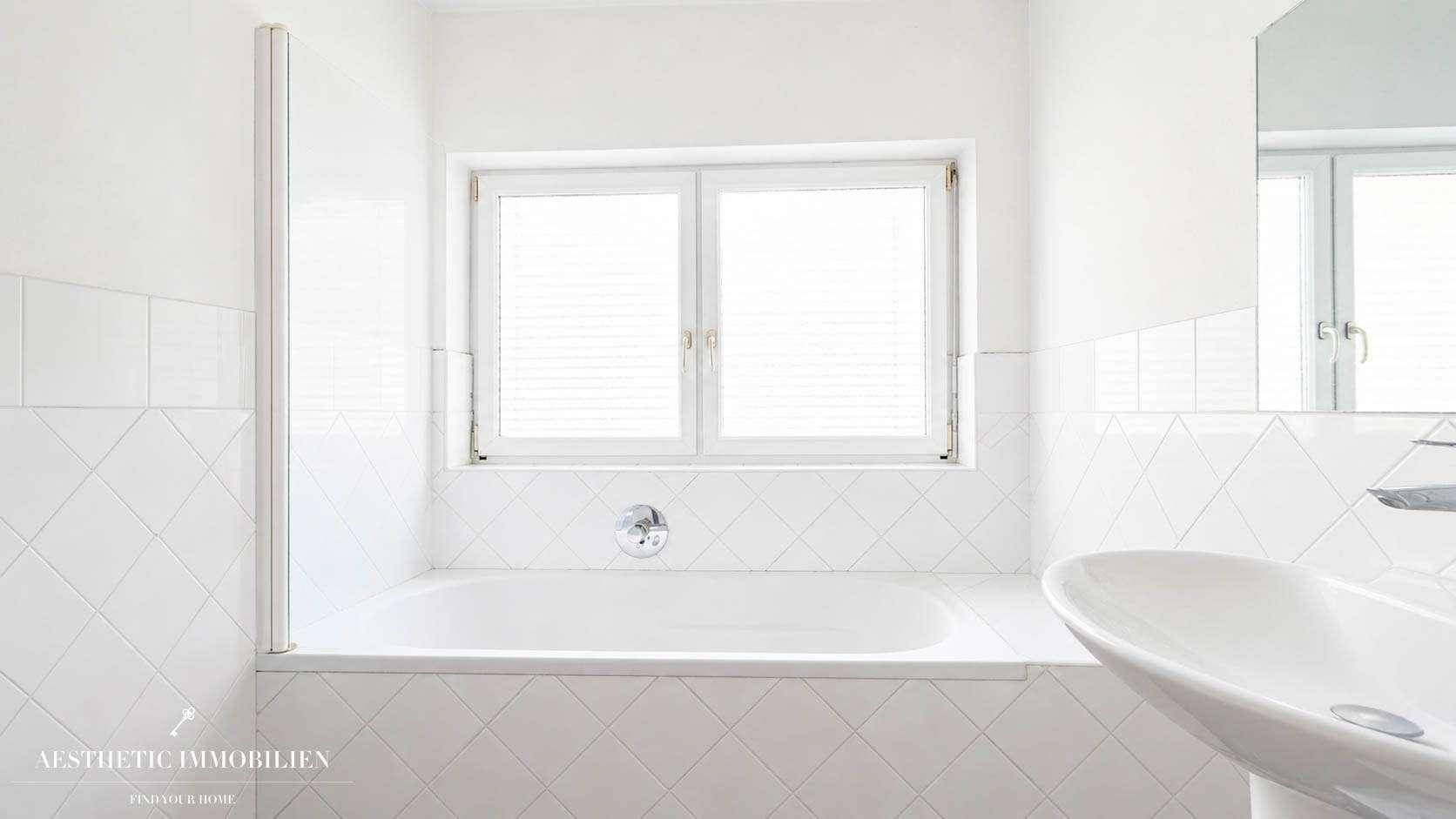 Helles Badezimmer mit eingebauter Badewanne, Waschbecken und Fenster, komplett gefliest.