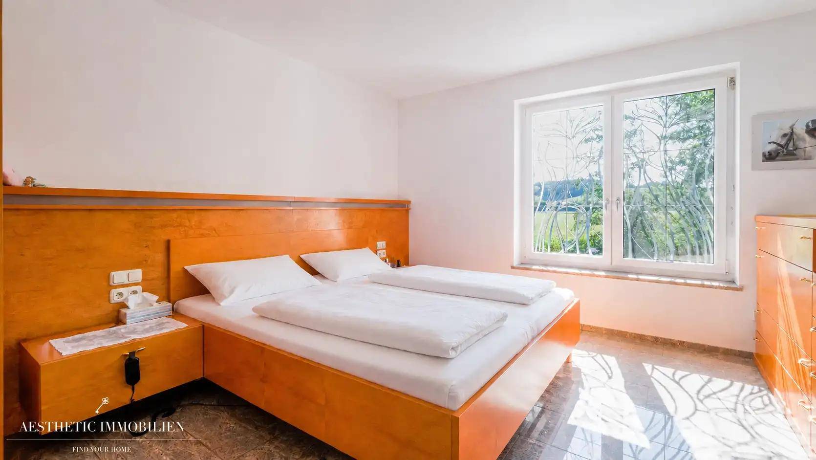 Schlafzimmer mit Doppelbett, Holzmöbeln und hellem Fenster mit Blick ins Grüne.