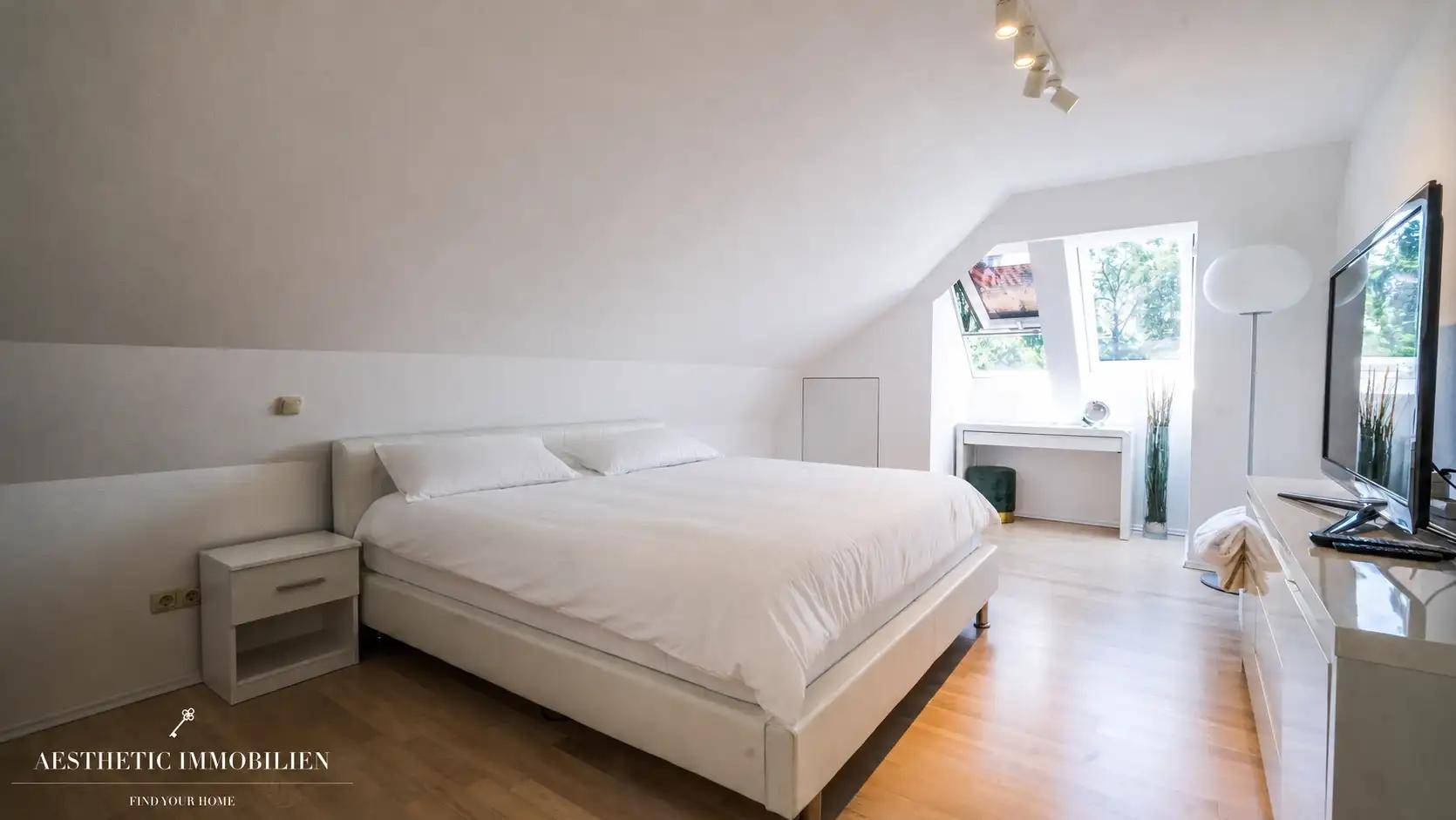Helles Schlafzimmer im Dachgeschoss mit weißem Bett, Holzboden und großem Dachfenster.