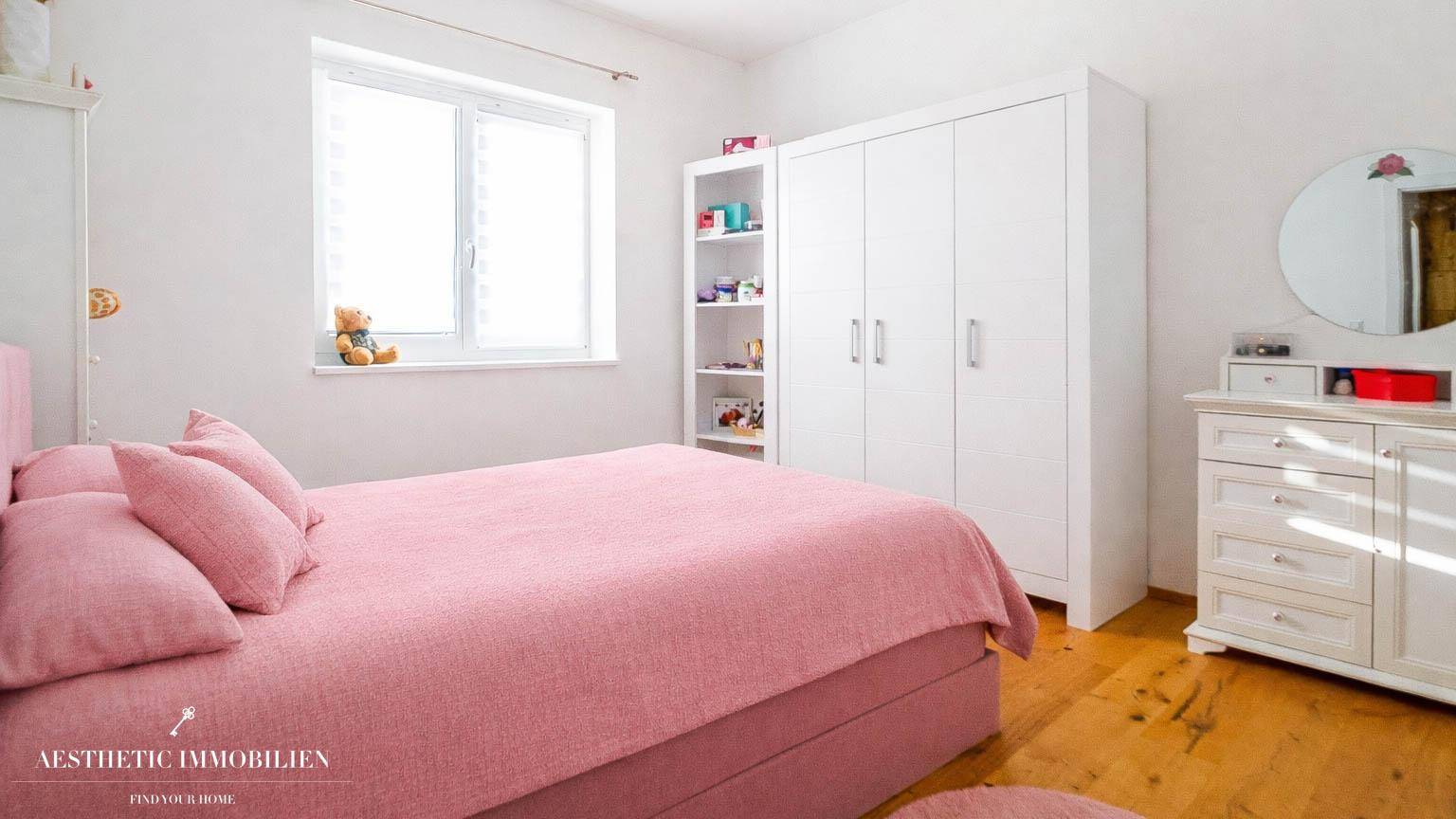 Kinderzimmer mit rosa Bett, weißem Kleiderschrank und großem Fenster.