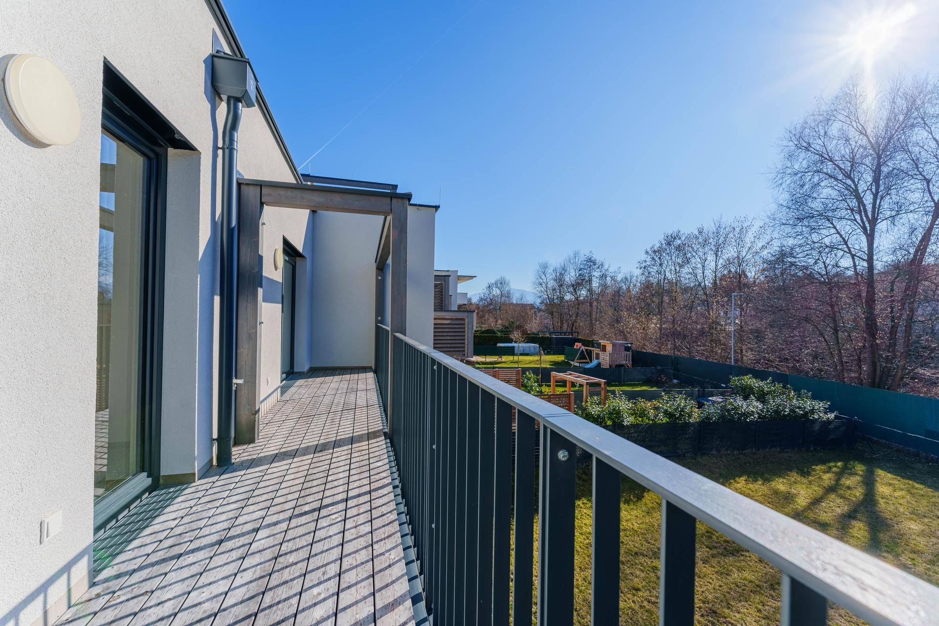 Sonniger Balkon mit Holzboden und Blick auf den Garten sowie einen kleinen Spielplatz.