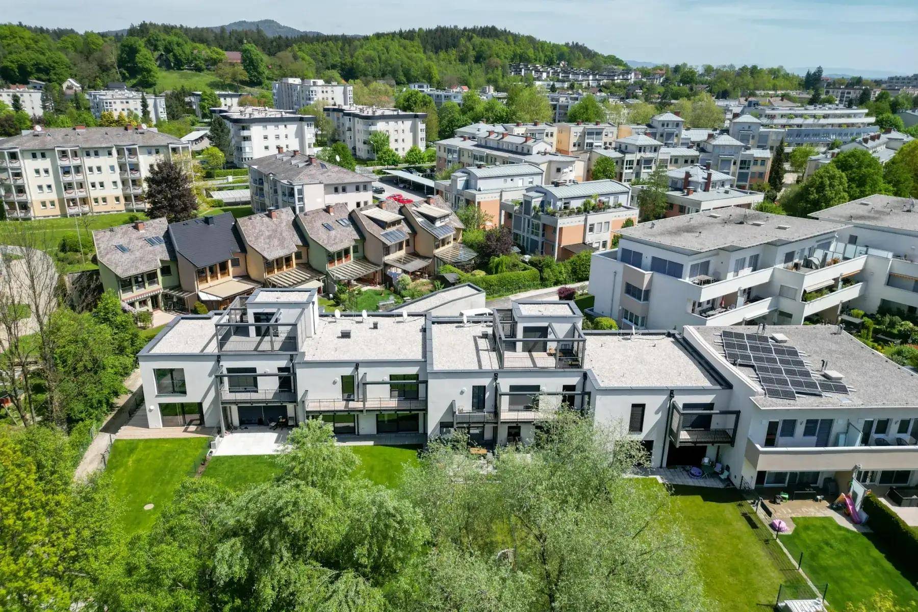 Luftaufnahme einer modernen Wohnsiedlung mit Reihenhäusern und Mehrfamilienhäusern, umgeben von viel Grün und Hügeln.
