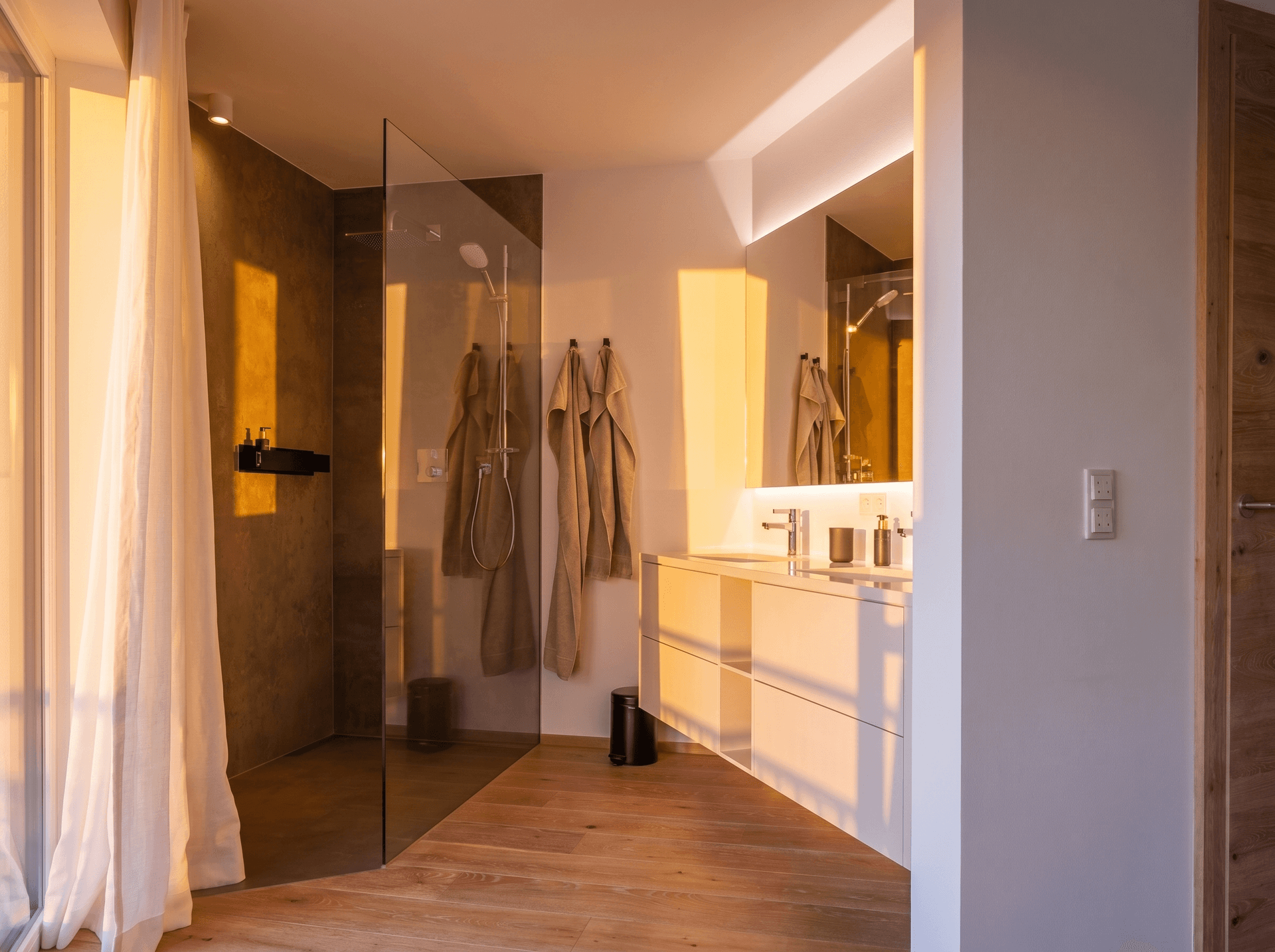 Modernes Badezimmer mit begehbarer Dusche, Doppelwaschbecken und Holzboden, durchflutet von warmem Abendlicht.