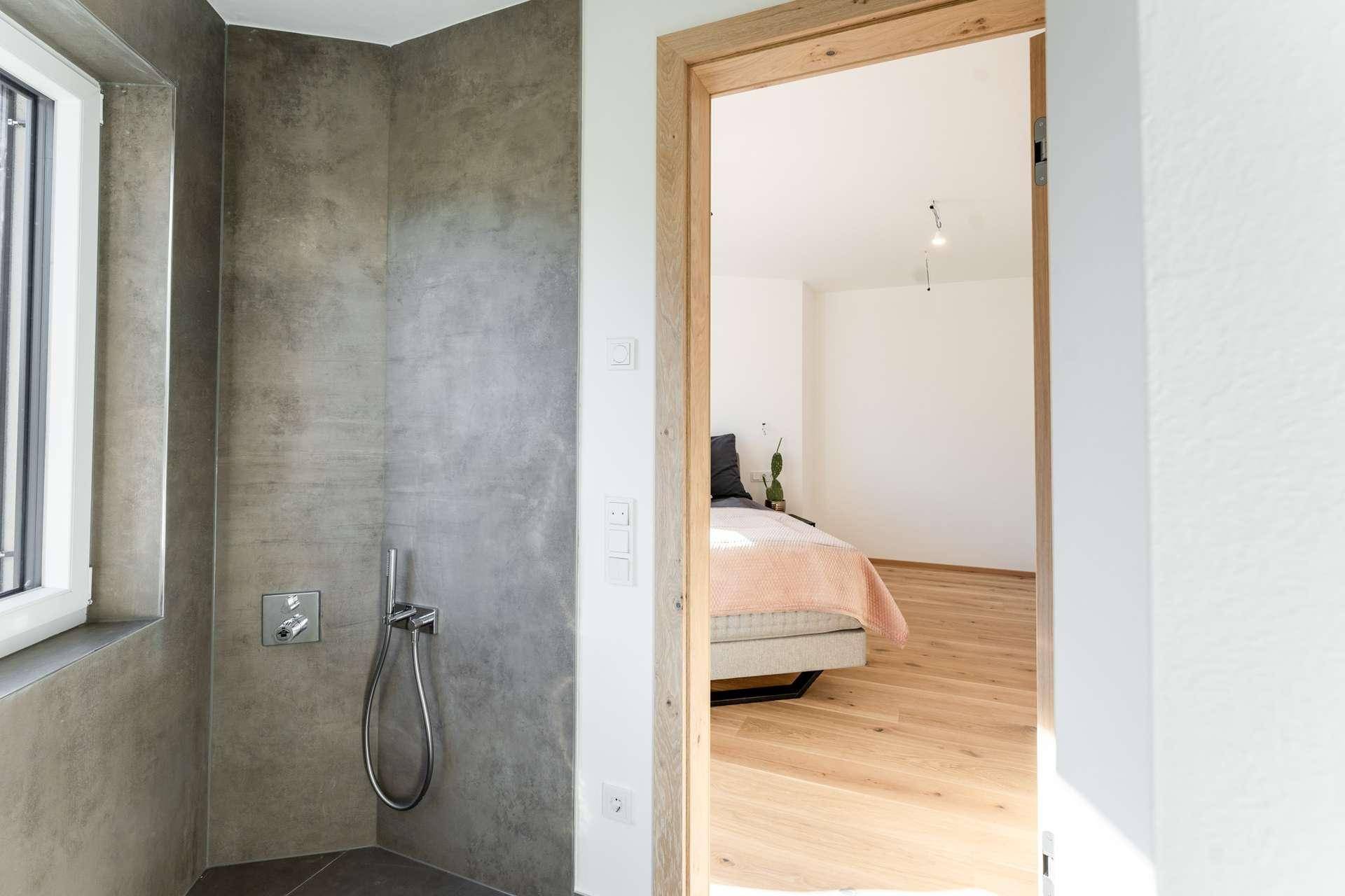 Modernes Badezimmer mit grauen Fliesen und Blick in ein helles Schlafzimmer mit Holzboden.