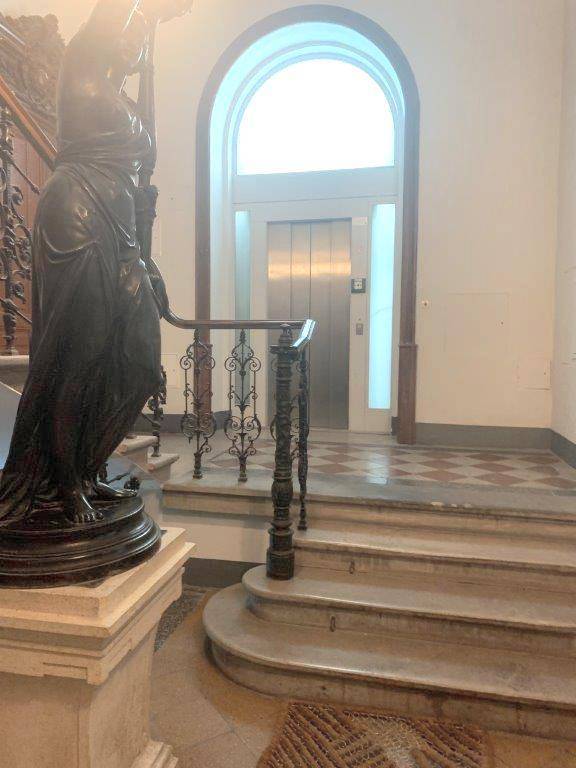 Prunkvolles Treppenhaus mit Statue, historischen Fliesen und modernem Aufzug im Hintergrund.