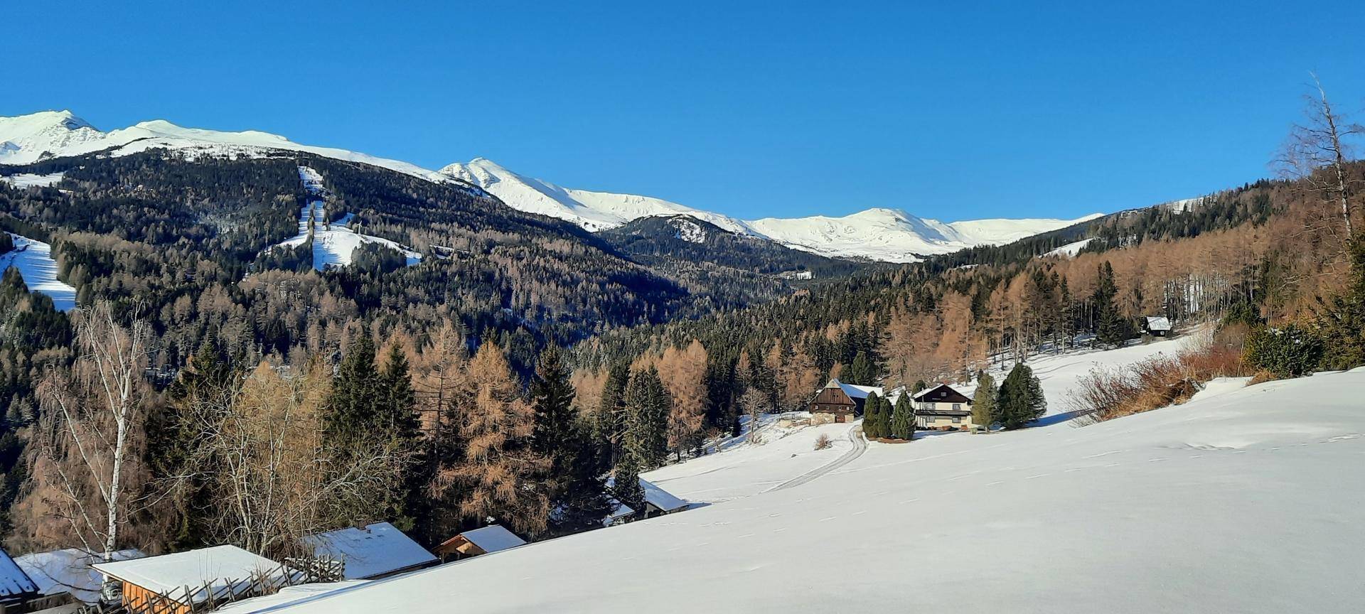 Weitläufige Winterlandschaft mit schneebedeckten Bergen, Wäldern und vereinzelten Häusern unter blauem Himmel.