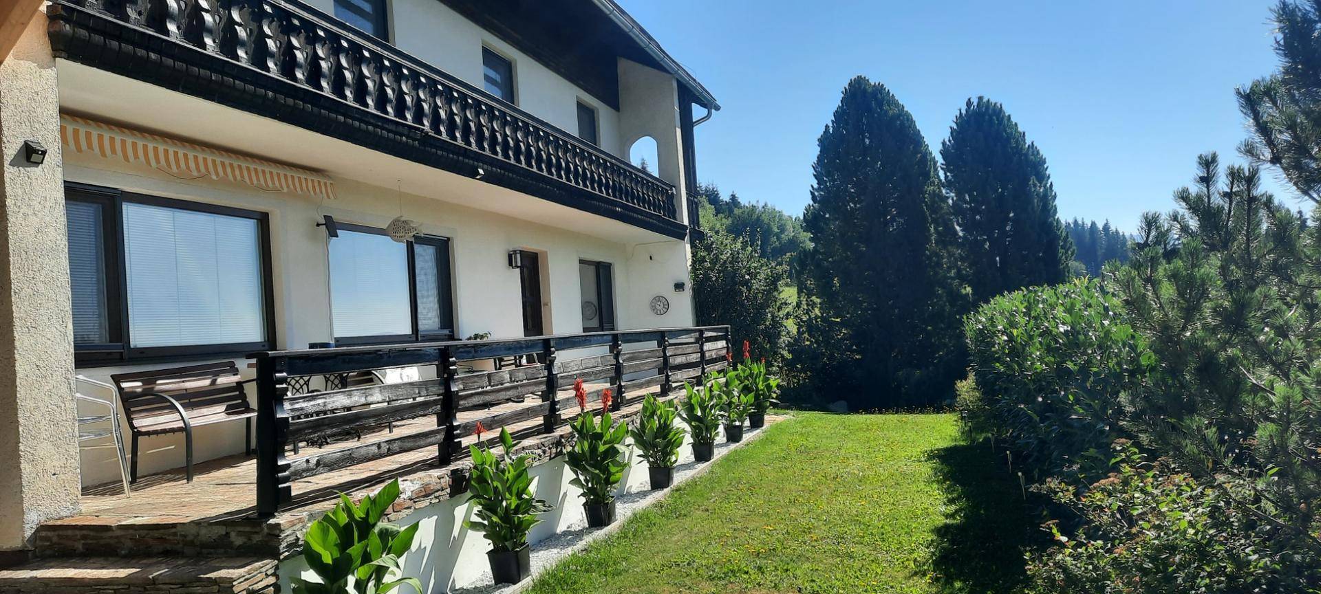 Helle Hausfassade mit einer einladenden Terrasse, einem Balkon und einem gepflegten Gartenbereich mit vielen Bäumen.