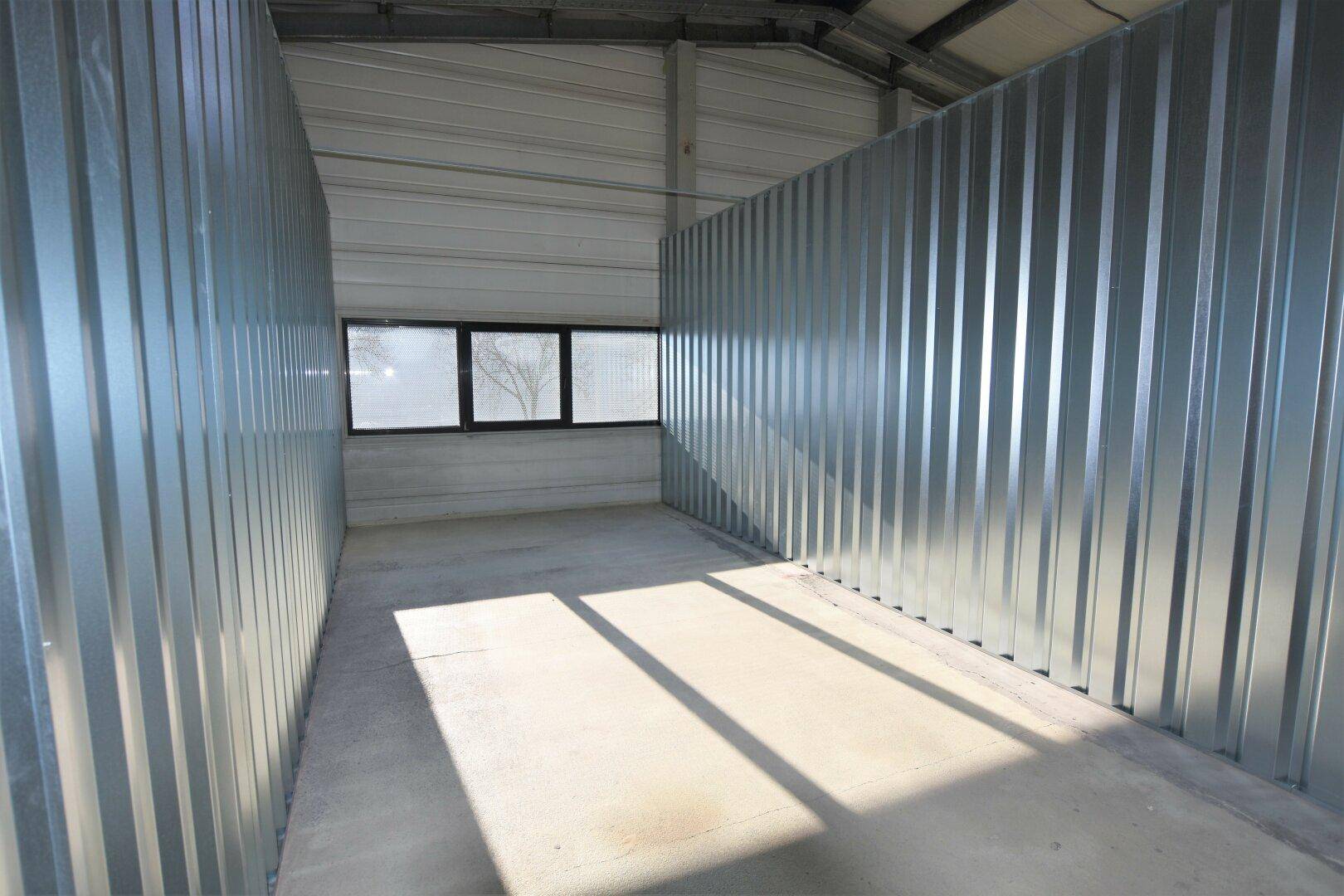 Helle Lagereinheit mit Wellblechwänden, Betonboden und einem Fenster, das Tageslicht spendet.