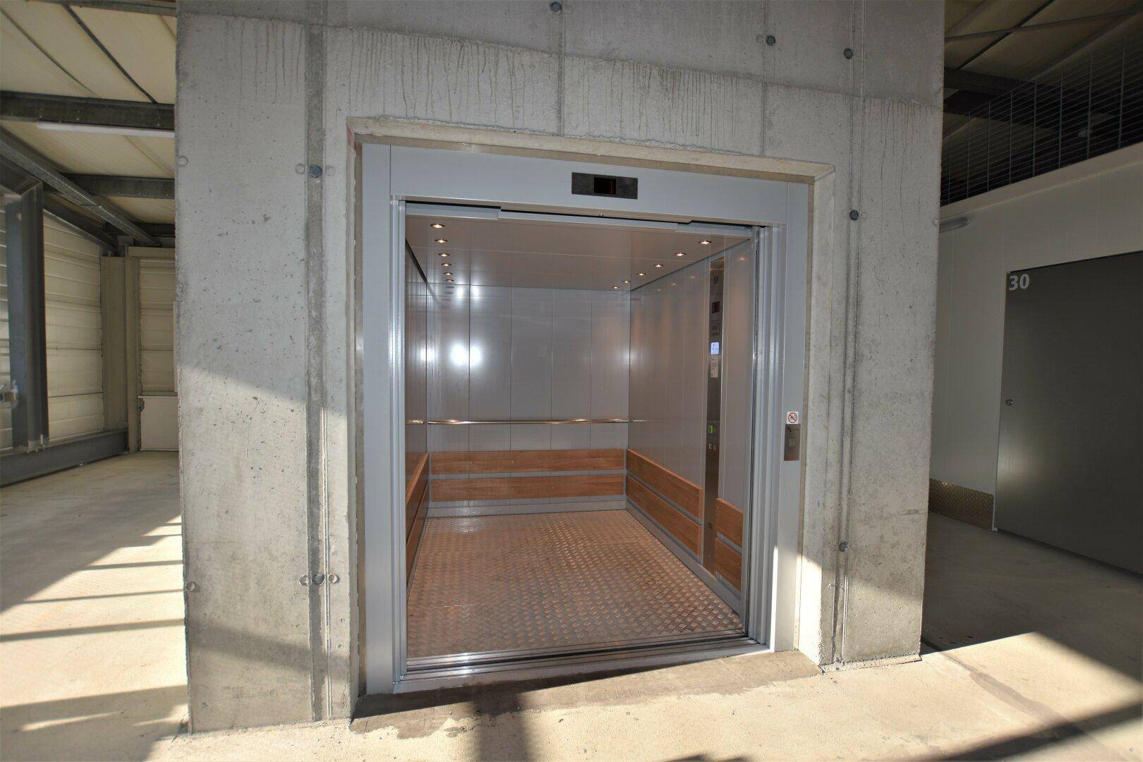 Moderner Aufzug in einem Betongebäude mit glänzenden Metallwänden und Holzakzenten im Inneren.
