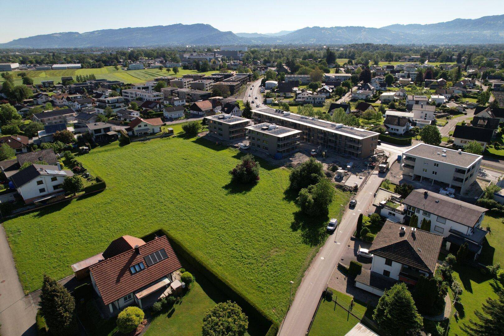 Panoramablick aus der Luft auf ein großes grünes Grundstück, umgeben von Wohngebäuden und einer Bergkette.