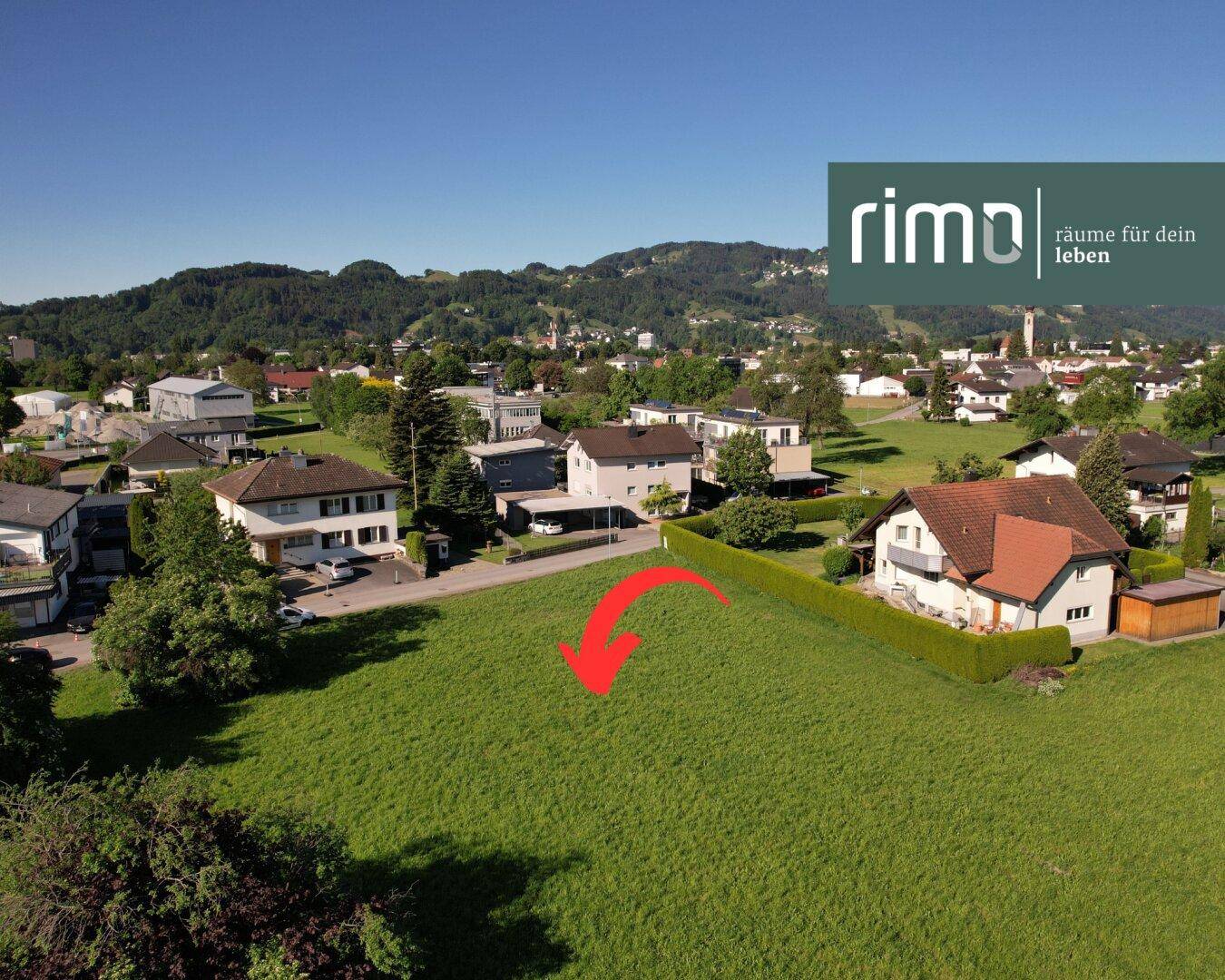 Weitläufiges grünes Grundstück aus der Vogelperspektive mit angrenzenden Wohnhäusern und malerischem Bergpanorama.