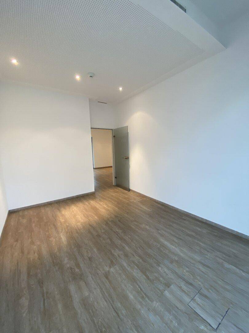 Leerer Raum mit hellem Holzboden, weißen Wänden und einer modernen Glastür zu einem weiteren Bereich.