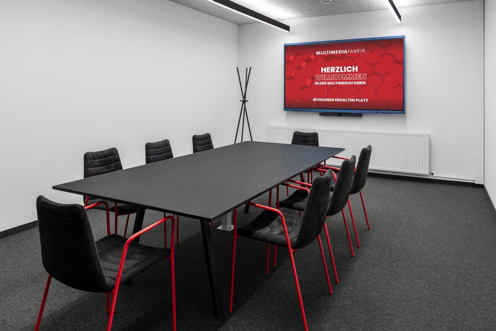 Moderner Konferenzraum mit großem Tisch, schwarzen Stühlen und einem Wandbildschirm.