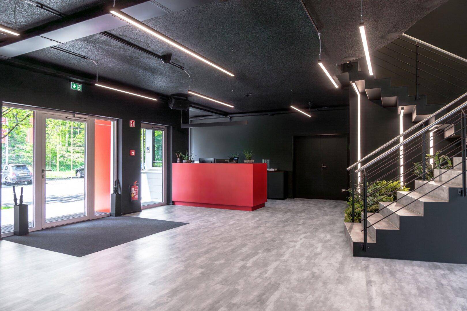 Heller Eingangsbereich mit roter Rezeptionstheke, großen Glastüren und einer modernen Treppe.