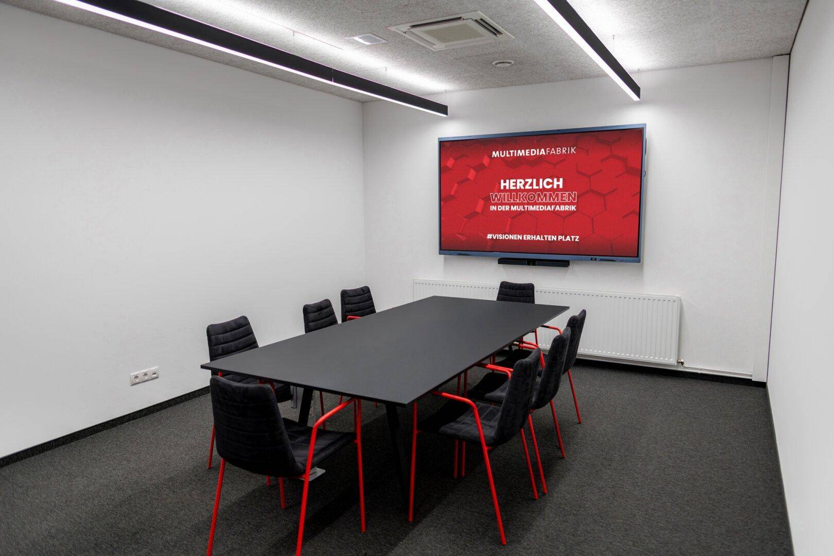 Heller Konferenzraum mit großem Tisch, schwarzen Stühlen und einem Wandbildschirm.