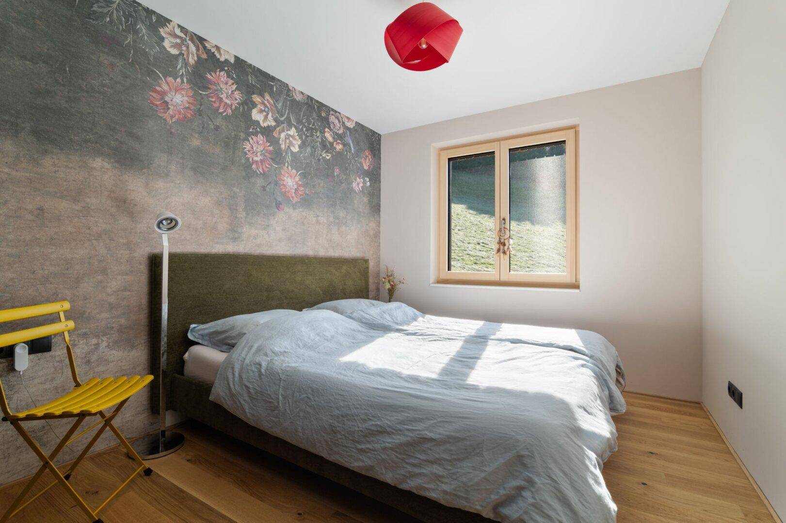 Helles Schlafzimmer mit Holzboden, floraler Tapete und Fenster mit Blick ins Grüne.