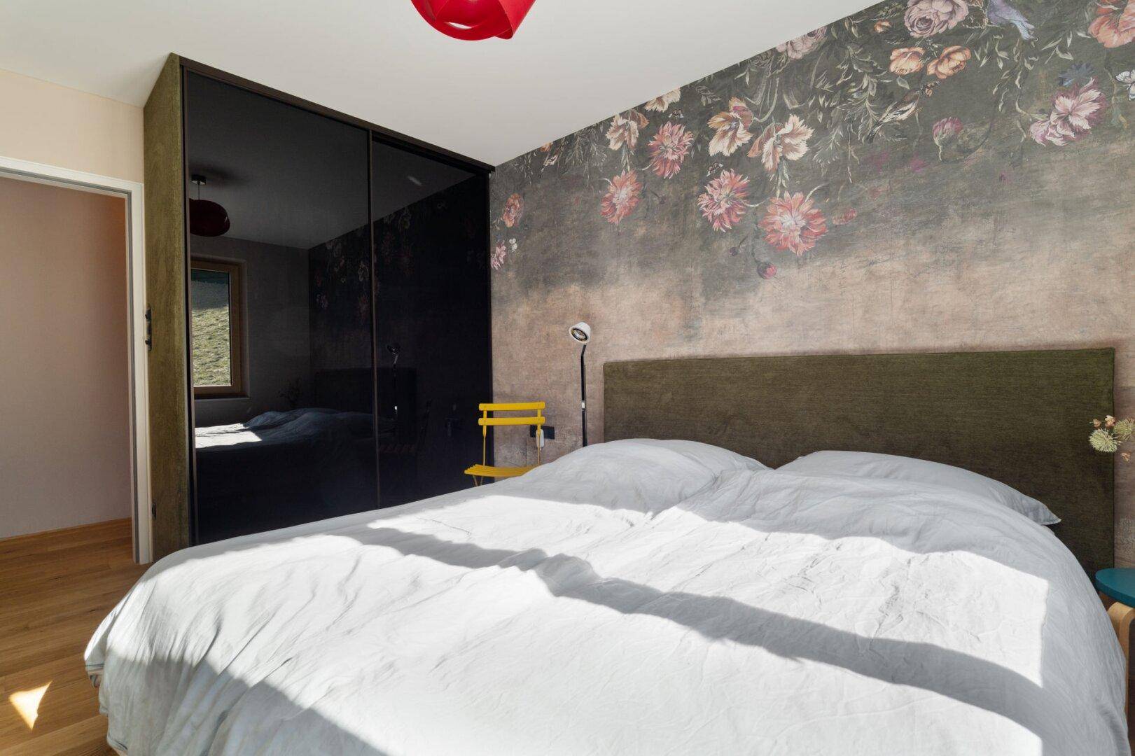 Schlafzimmer mit floraler Akzentwand, Holzboden und großem Spiegelschrank.