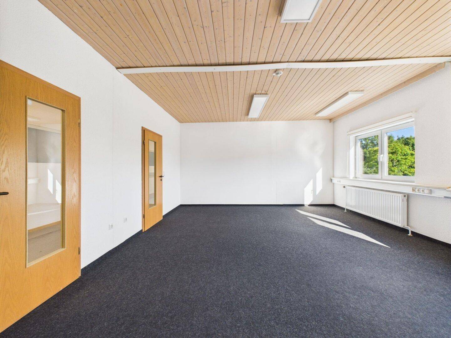 Geräumiger Büroraum mit Teppichboden, Holzlattendecke und Fenstern, die viel natürliches Licht hereinlassen.