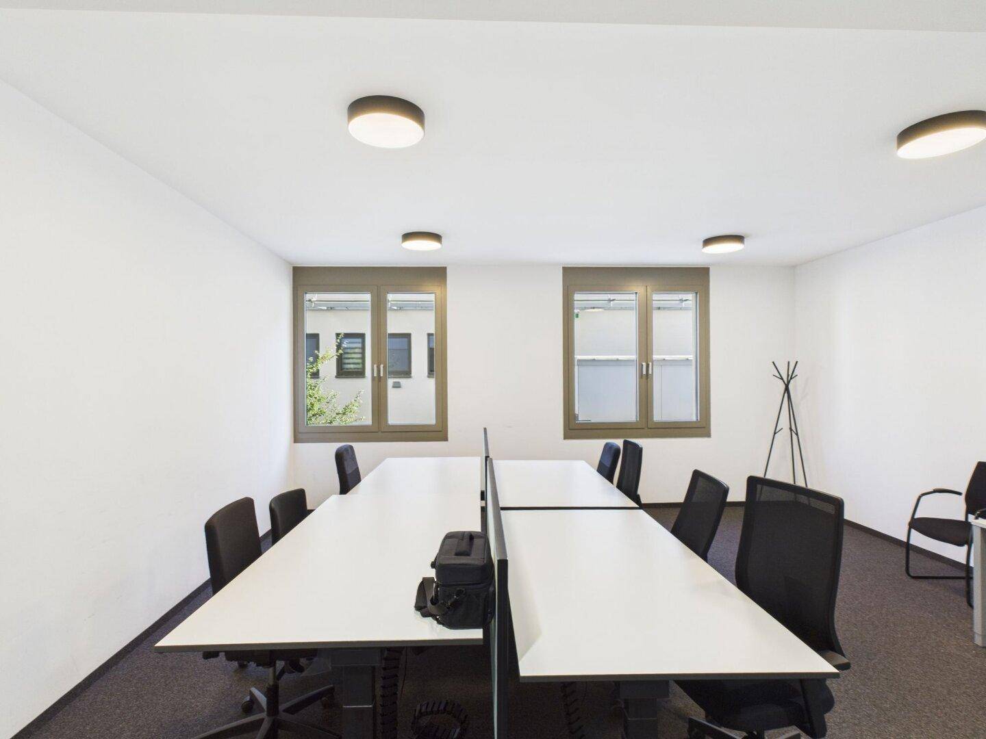 Moderner Büroraum mit mehreren Schreibtischen, ergonomischen Stühlen und großen Fenstern für Tageslicht.
