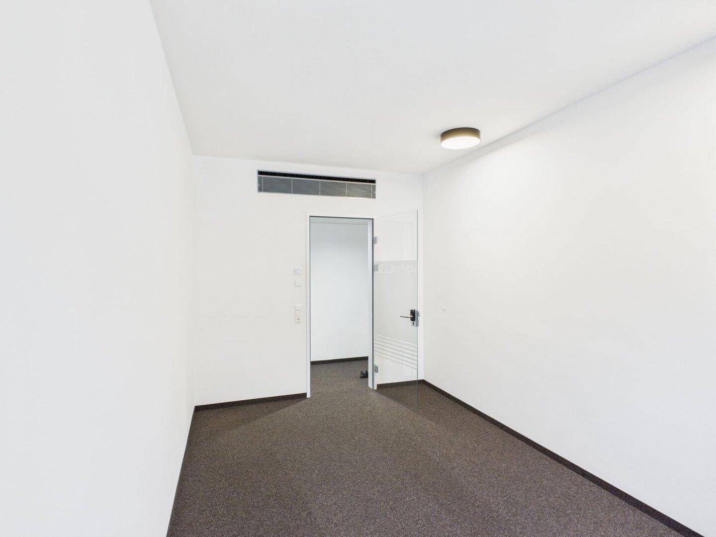 Leerer Büroraum mit weißen Wänden, Teppichboden und einer Lüftungsanlage über der Tür.