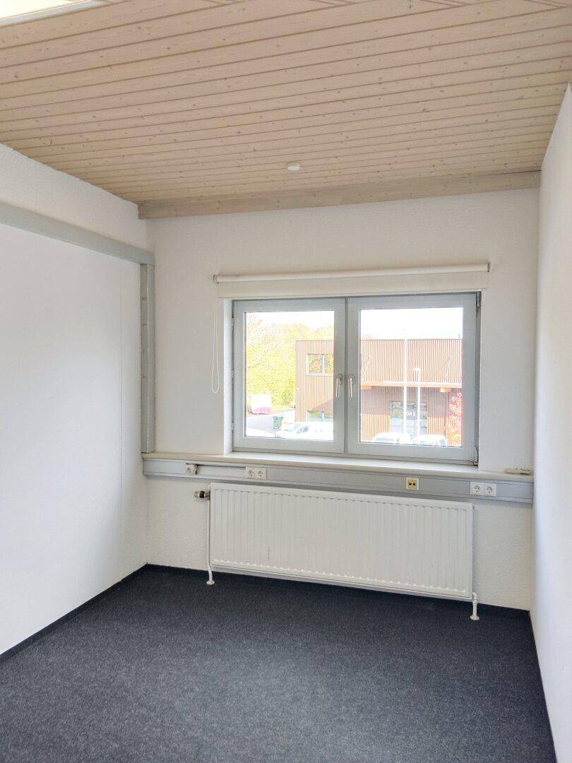 Kleiner, heller Büroraum mit Teppichboden, Holzdecke und einem Fenster, das Tageslicht hereinlässt.
