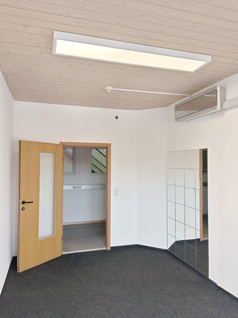 Kleiner Büroraum mit Teppichboden, Holzdecke und einer Holztür, die zu einem weiteren Raum führt.