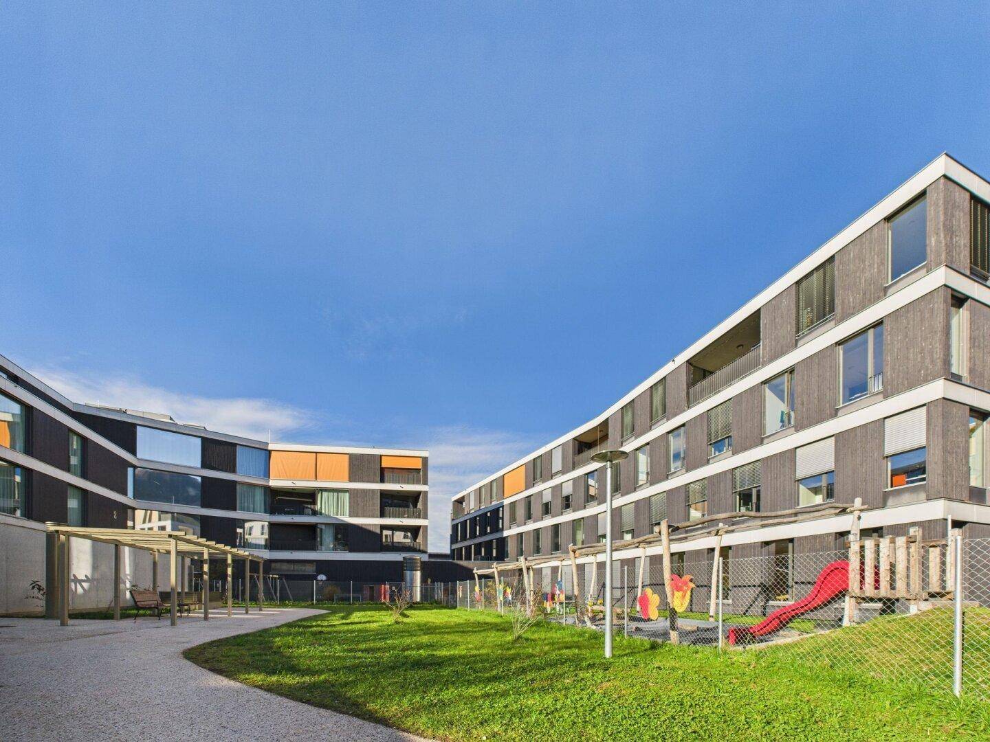 Innenhof eines modernen Gebäudekomplexes mit Spielplatz, Rasenfläche und Sitzgelegenheiten.