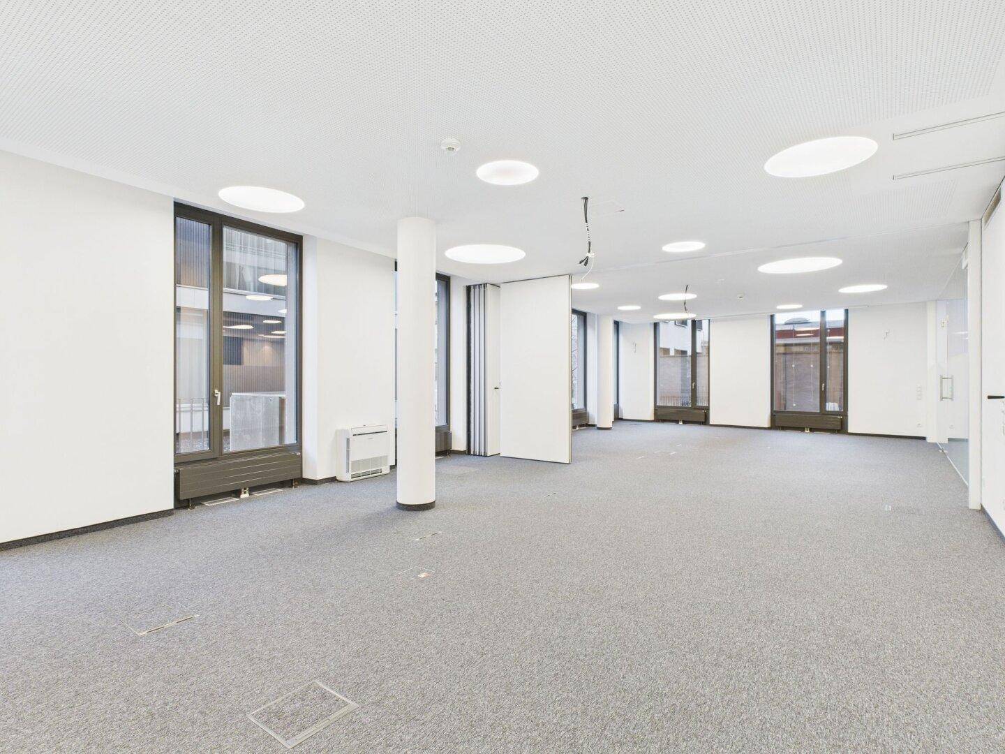 Heller, offener Büroraum mit Teppichboden, vielen Fenstern und moderner Deckenbeleuchtung.