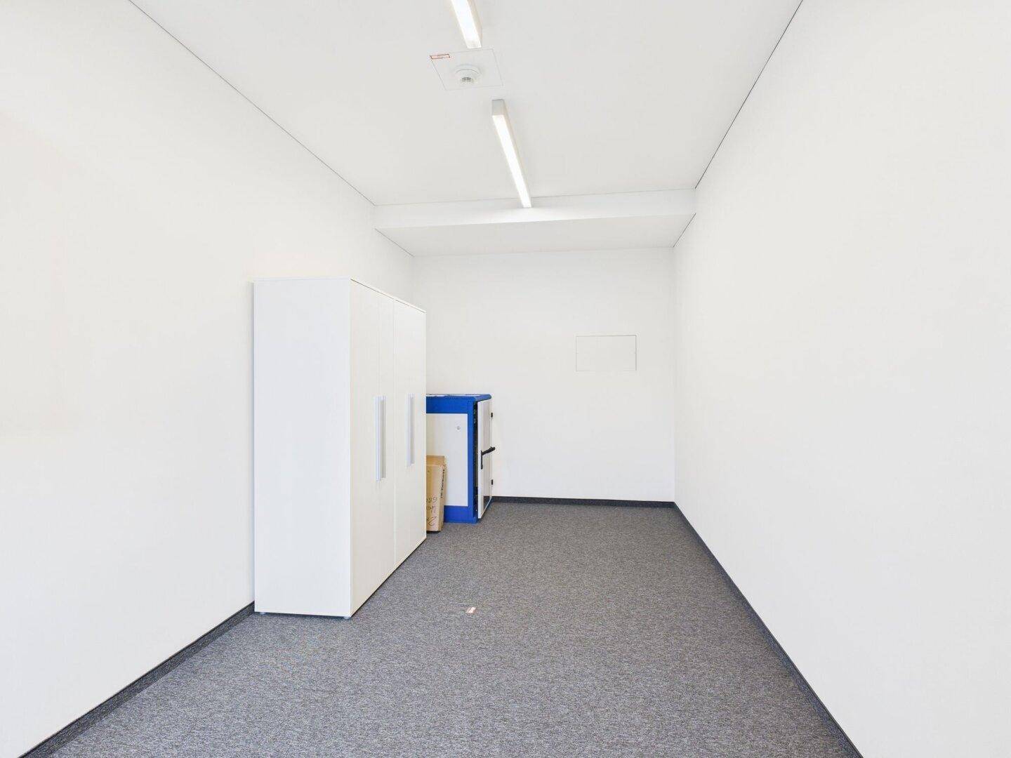 Schmaler Büroraum mit Teppichboden, weißen Wänden und praktischen Schränken für Stauraum.