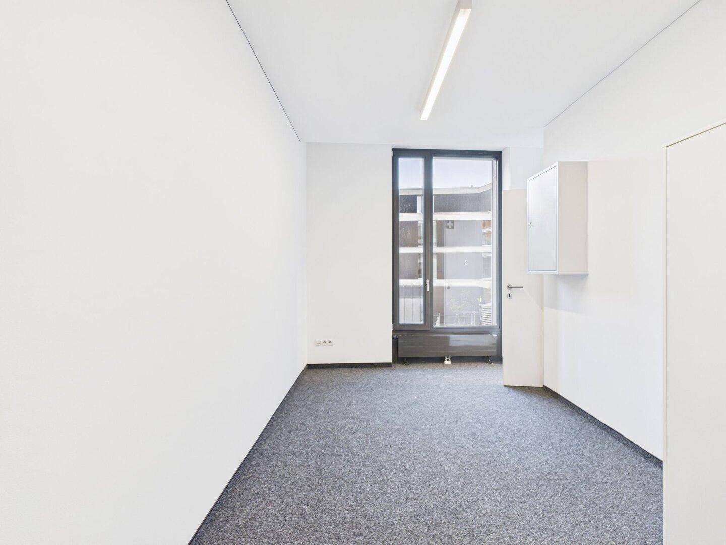 Kleiner, heller Büroraum mit Teppichboden, weiß gestrichenen Wänden und einem großen Fenster.