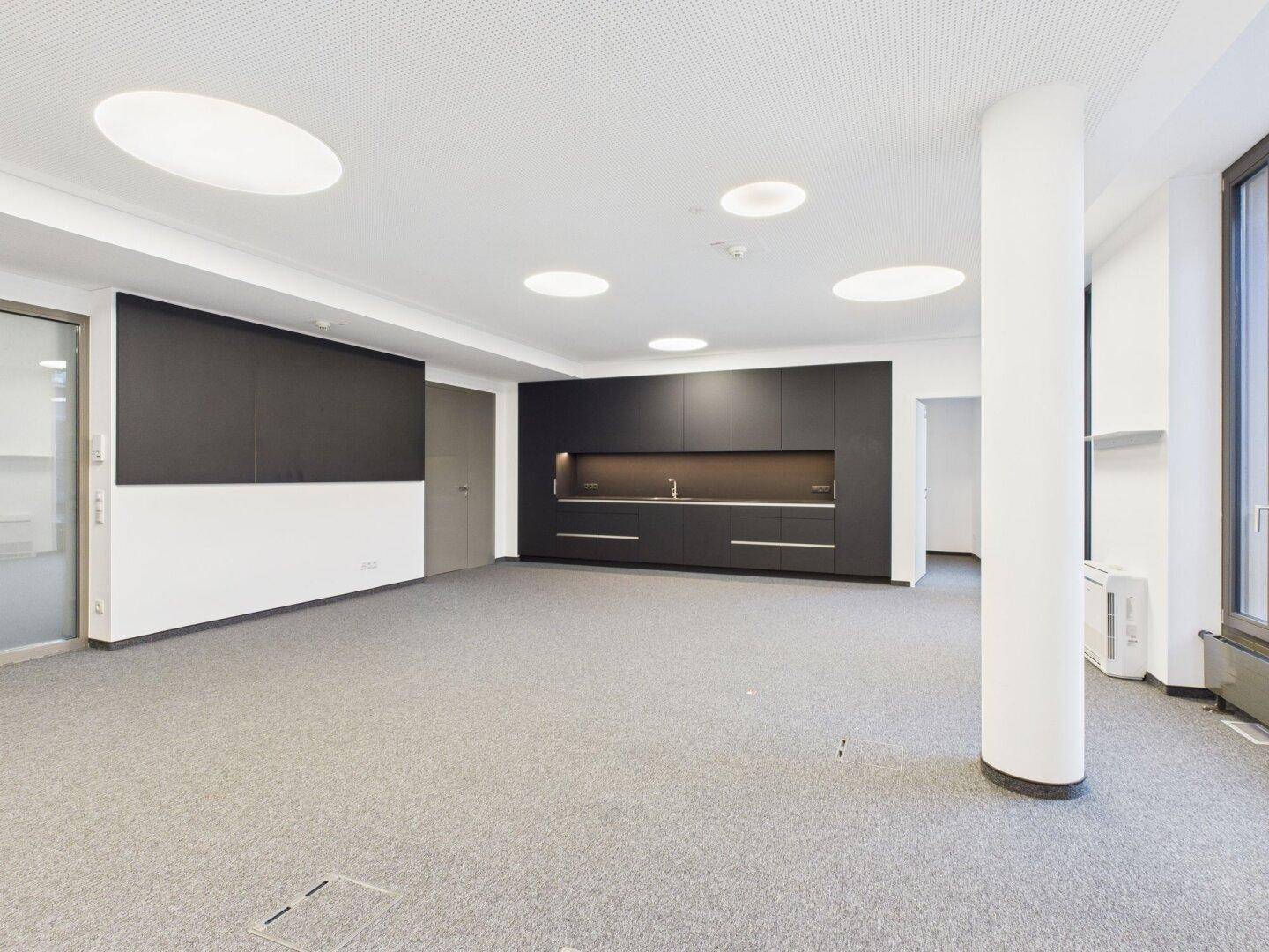 Geräumiger Bürobereich mit integrierter Küchenzeile, Säule und großen Fenstern für viel Tageslicht.