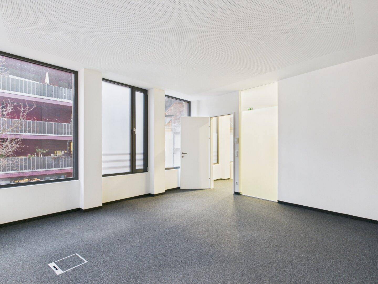 Geräumiges Büro mit großen Fenstern, grauem Teppichboden und viel Tageslicht.