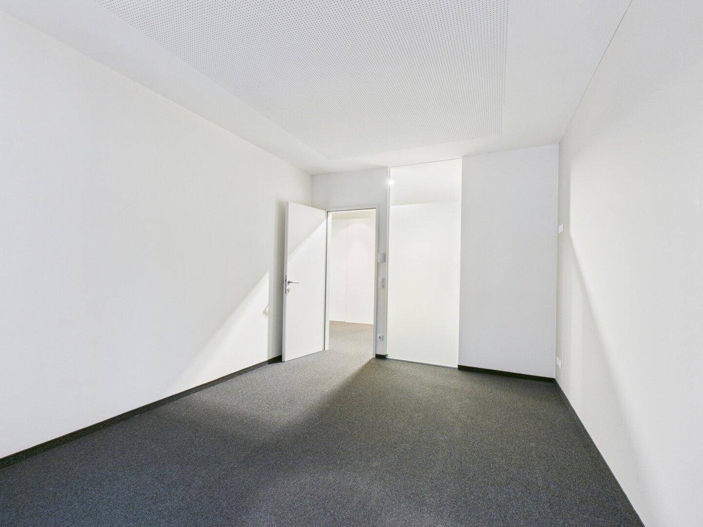 Leeres Büro mit grauem Teppichboden und einer Tür, die zu einem weiteren Raum führt.