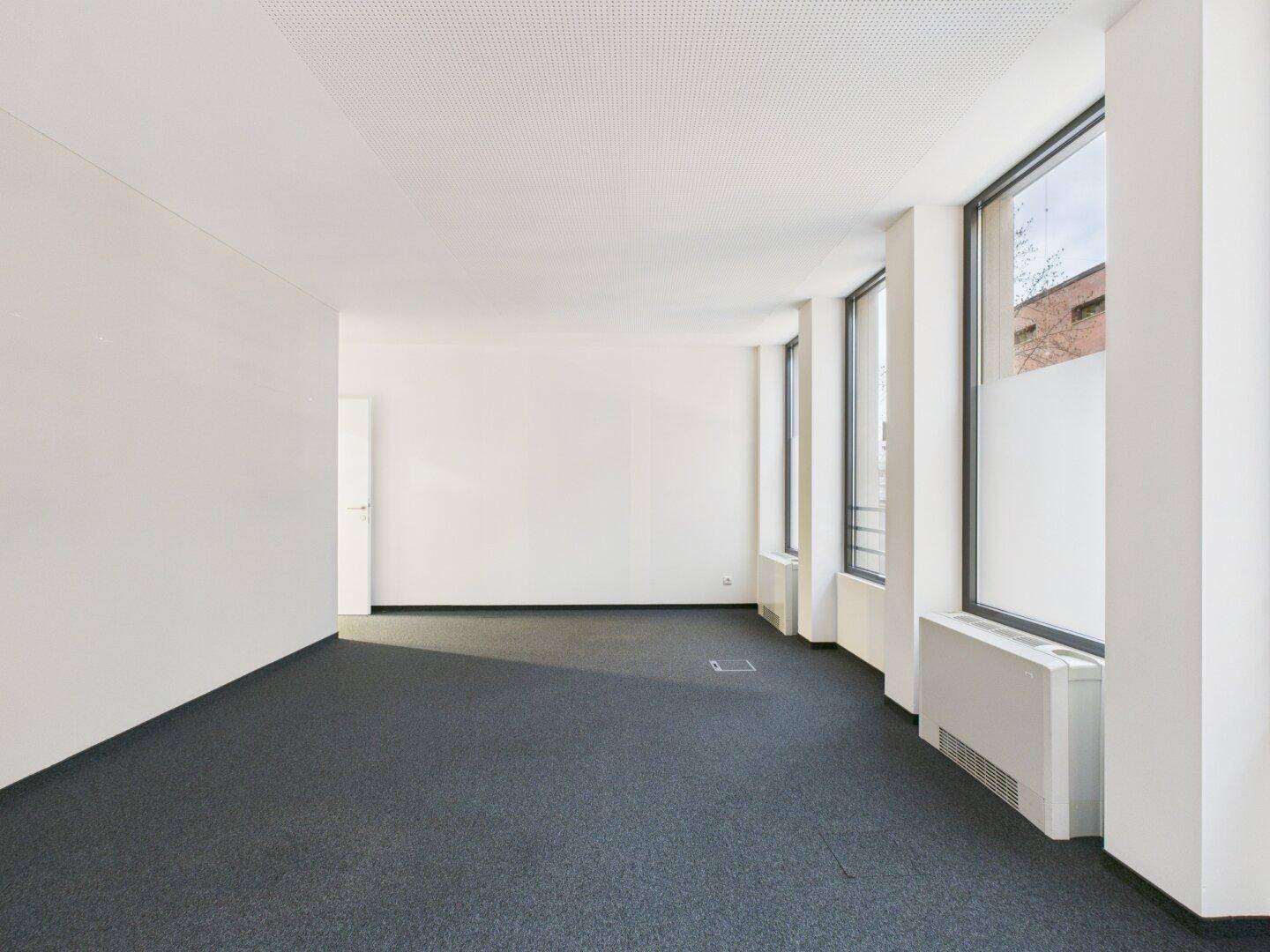 Langes, helles Büro mit Teppichboden und mehreren Fenstern, die für eine gute Belichtung sorgen.