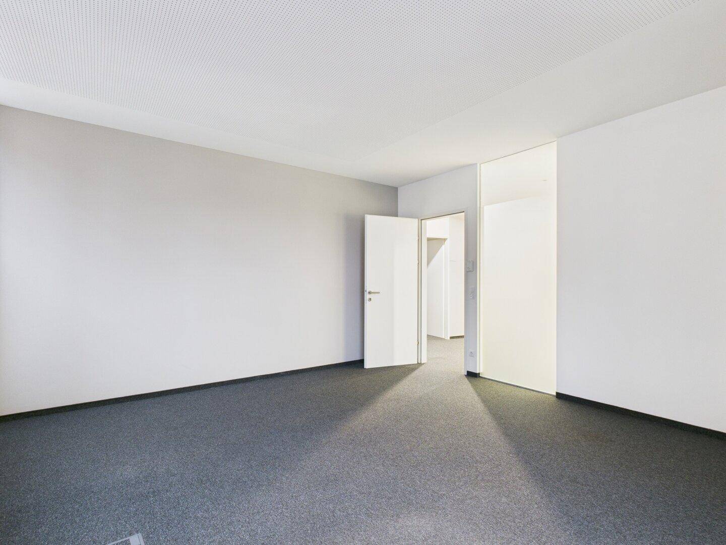 Helles und geräumiges Büro mit grauem Teppichboden und einer Tür, die zu einem weiteren Bereich führt.