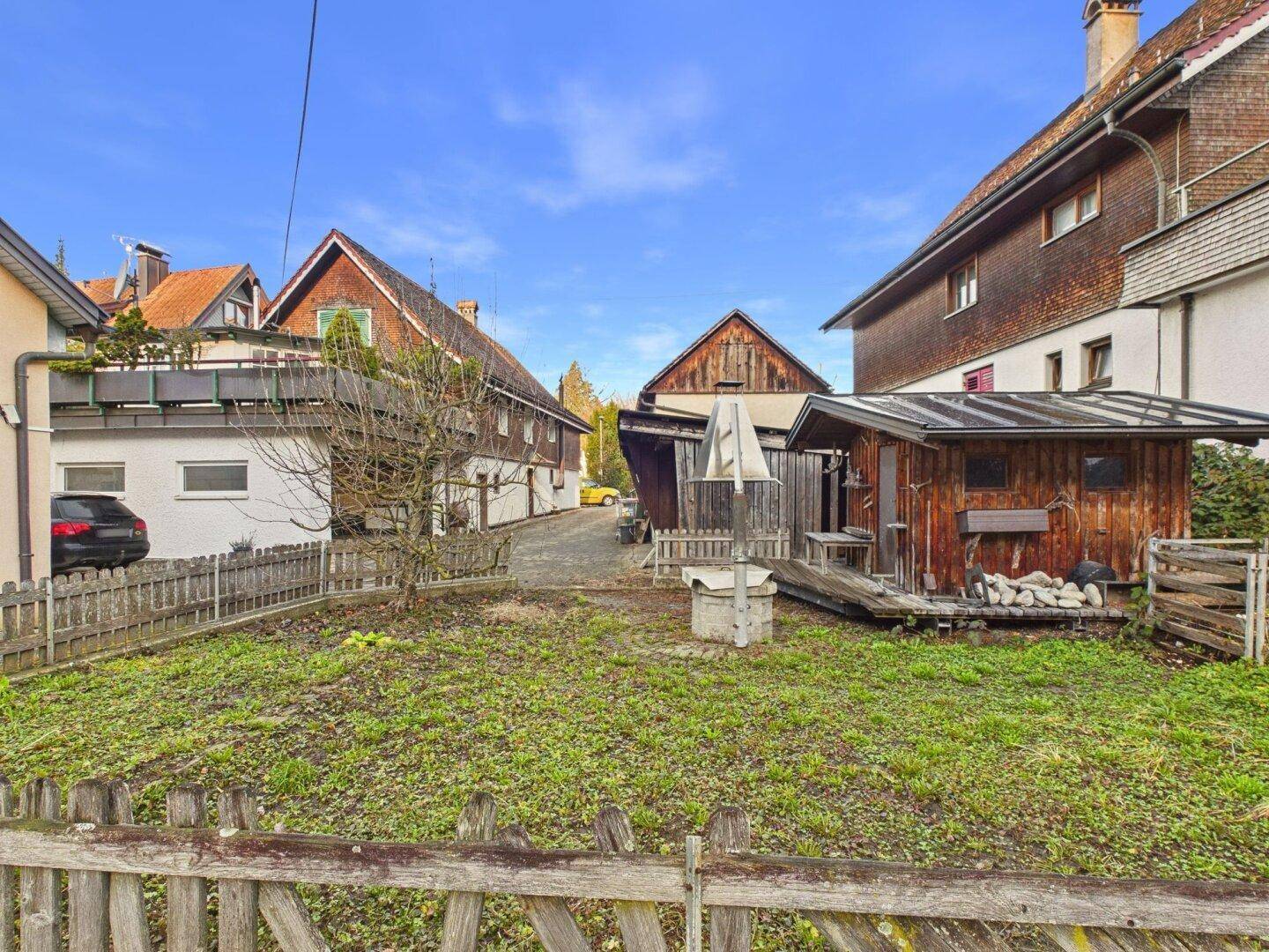 Grüne Gartenfläche mit Holzzaun und Blick auf die traditionellen Gebäude der Umgebung.