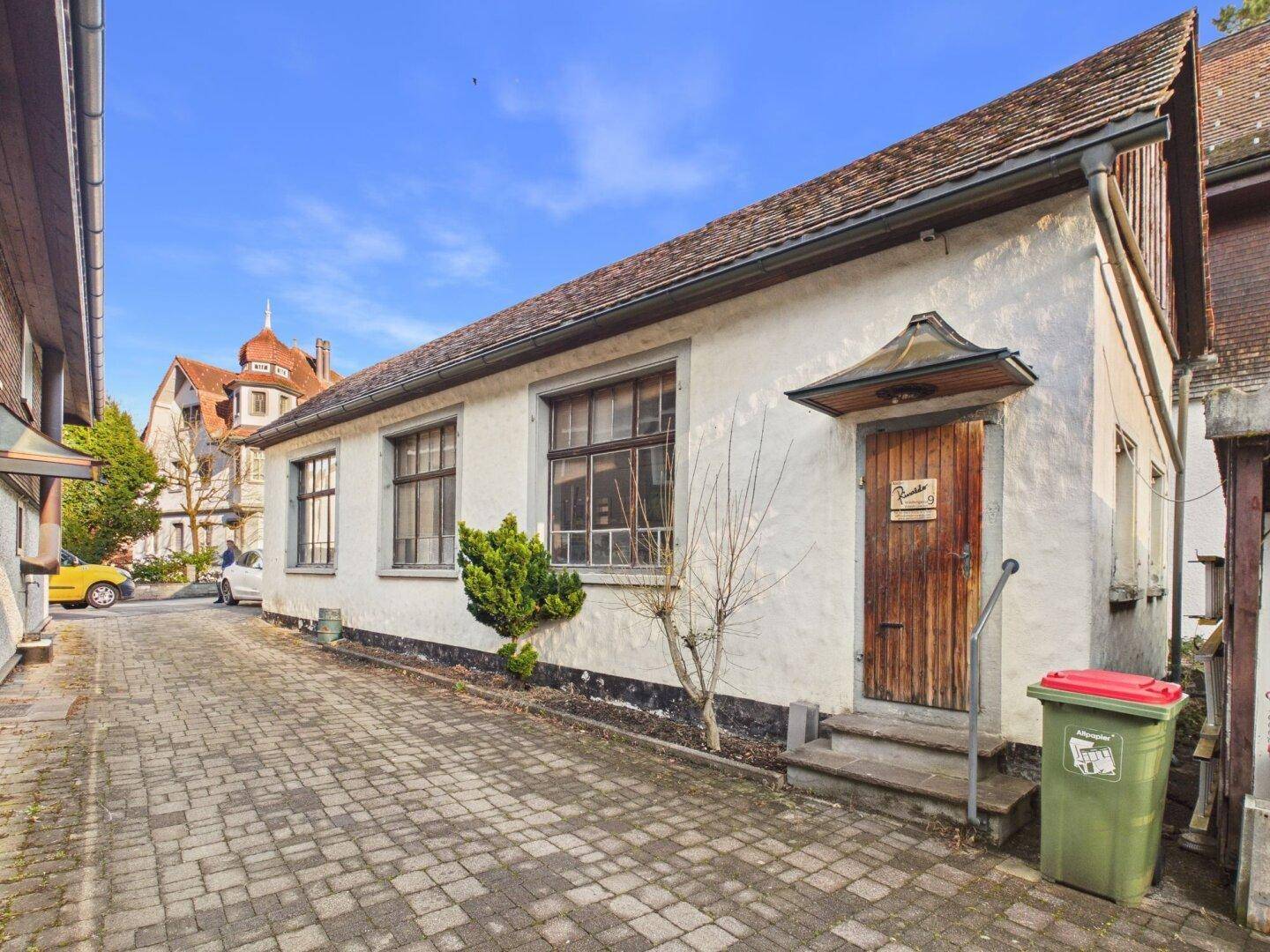 Traditionelles weißes Gebäude mit Holztür und Vordach, angrenzend an einen gepflasterten Weg.
