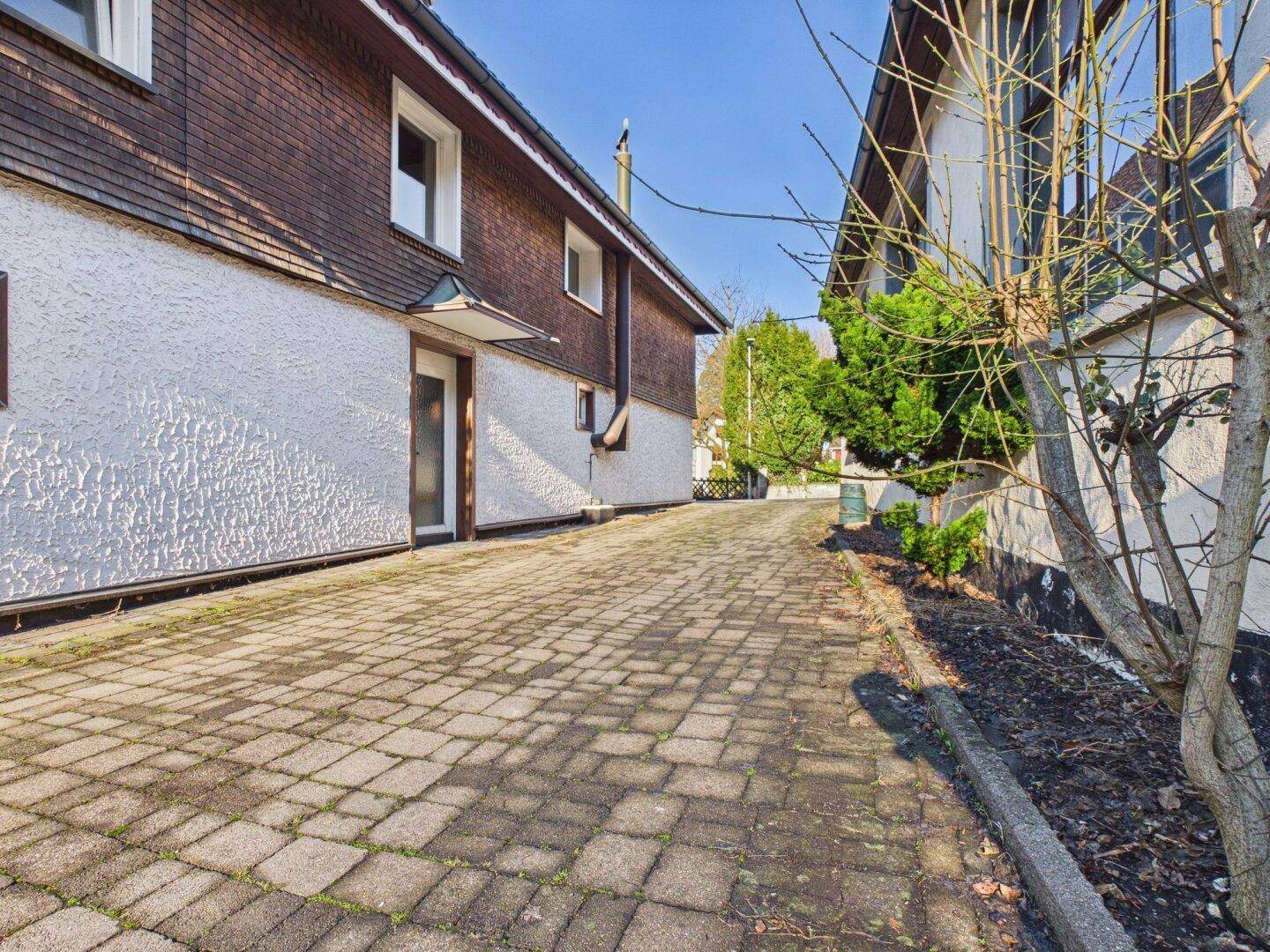 Gepflasterte Zufahrt zwischen zwei Gebäuden mit schattigen Bereichen und Baumbestand.