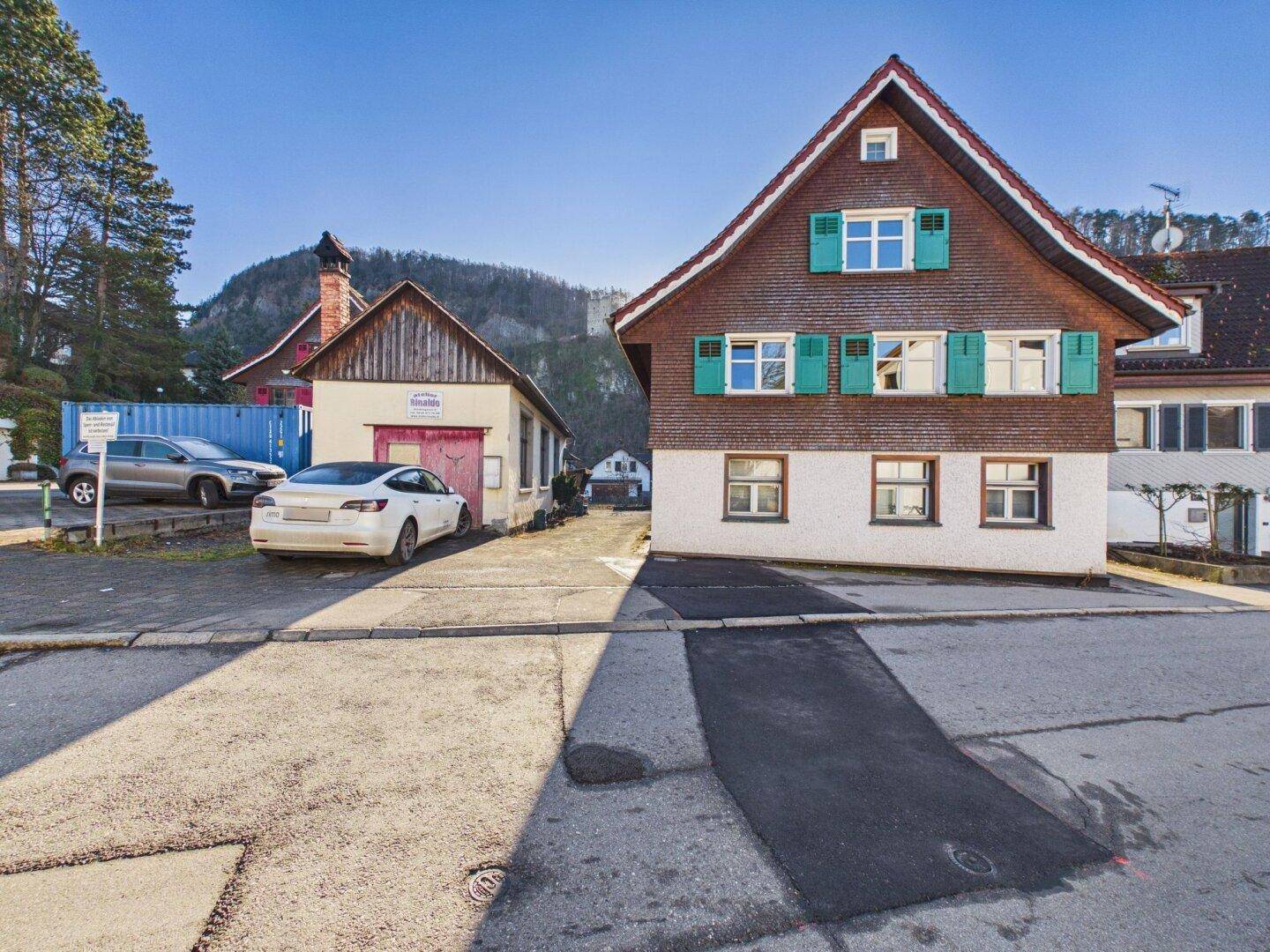 Traditionelles Haus mit grünen Fensterläden und Bergblick, umgeben von einer ländlichen Umgebung.