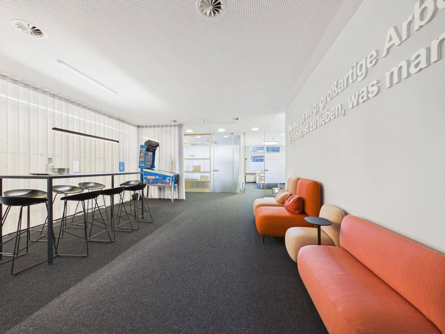 Entspannter Loungebereich mit modernen Sofas, Stehtischen und einem Flipperautomaten zur Unterhaltung.
