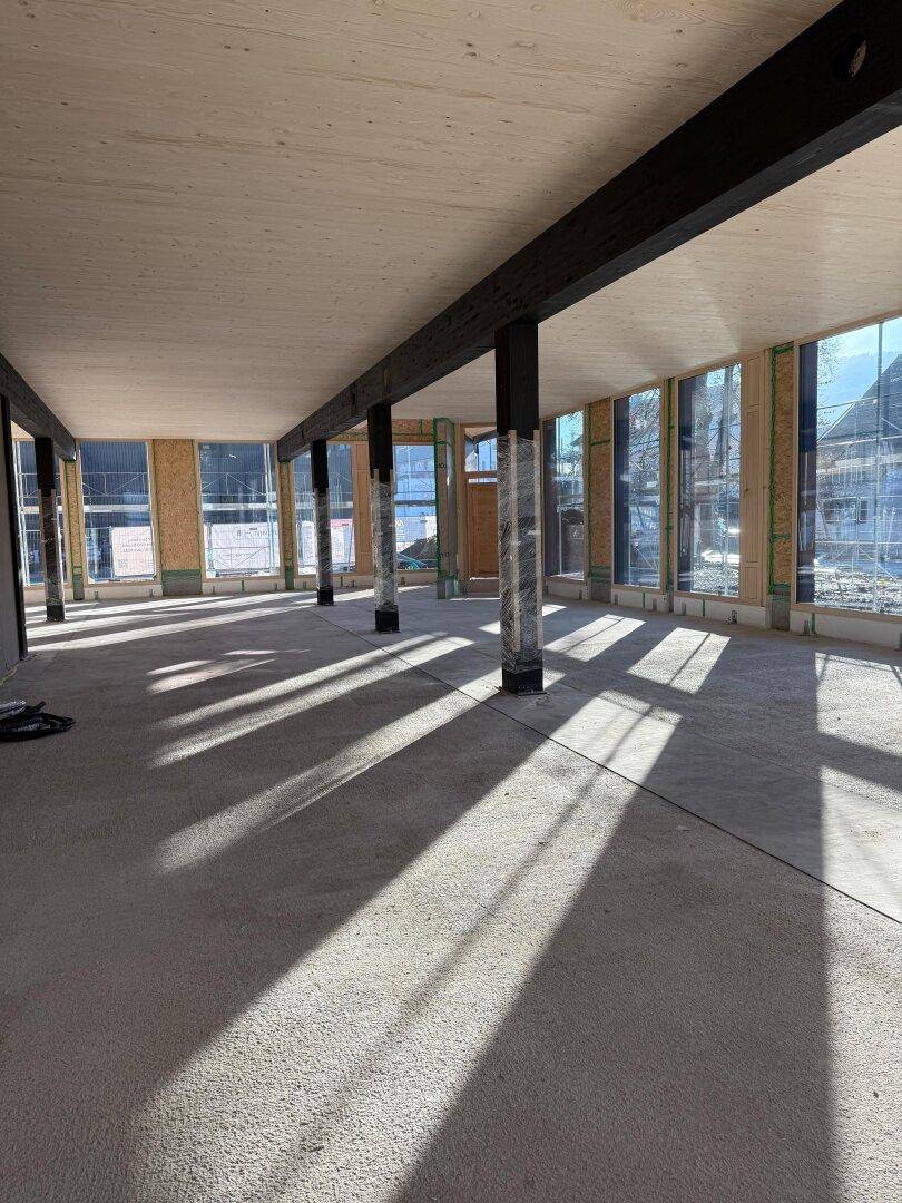 Geräumiger, unfertiger Bürobereich mit Betonboden, Holzbalken und viel natürlichem Lichteinfall durch große Fenster.