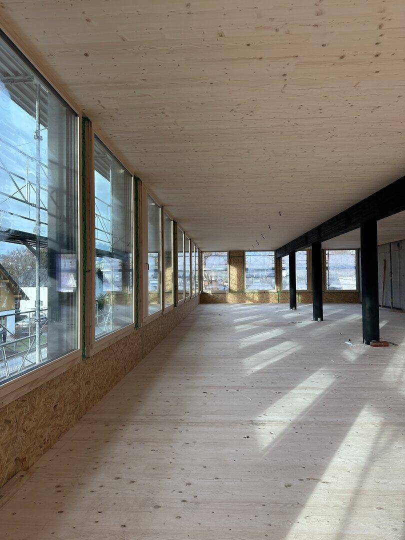 Langer, unfertiger Büroraum mit Holzboden und einer durchgehenden Fensterfront für viel Tageslicht.