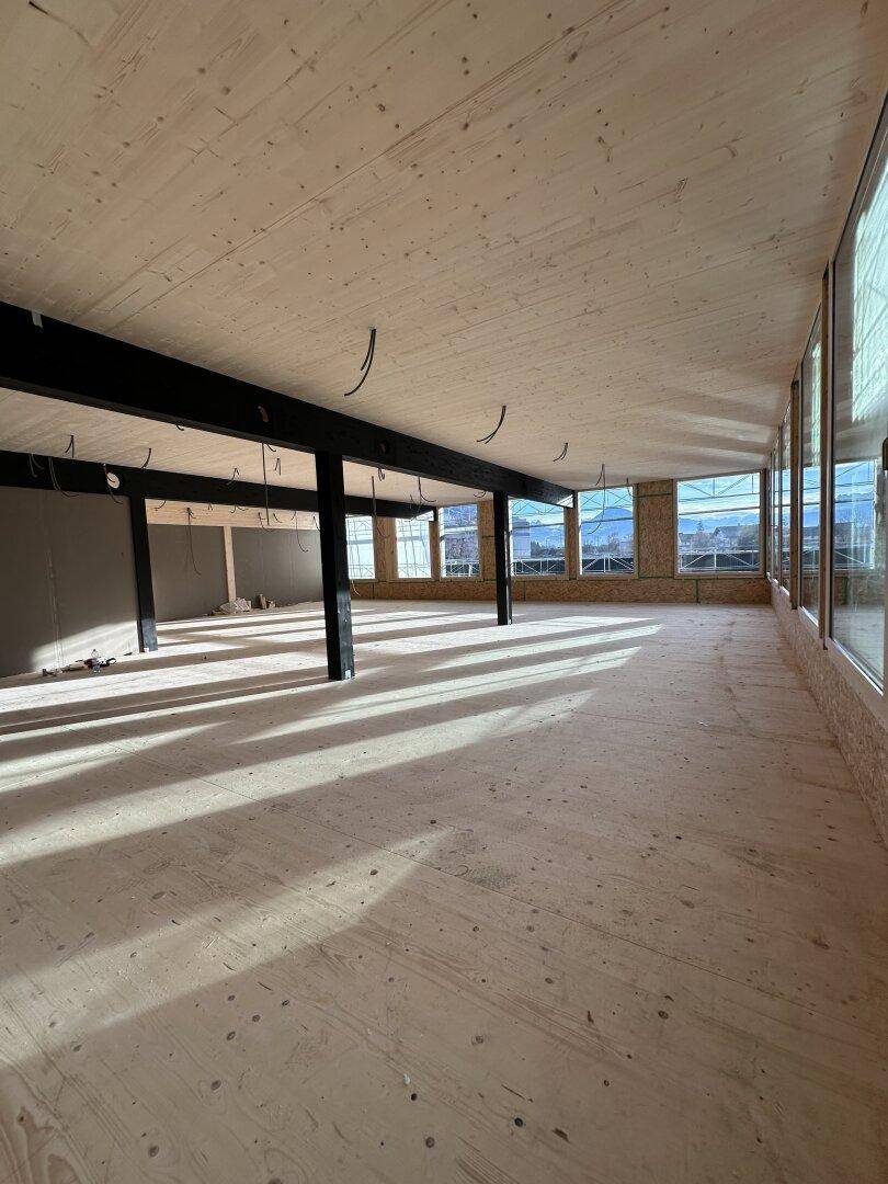 Heller, unfertiger Bürobereich mit Holzboden, Holzbalken und großen Fenstern, die viel Sonnenlicht hereinlassen.